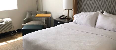 A hotel room with a large bed, a nightstand with a lamp, a chair, a desk, and a wall-mounted air conditioner.