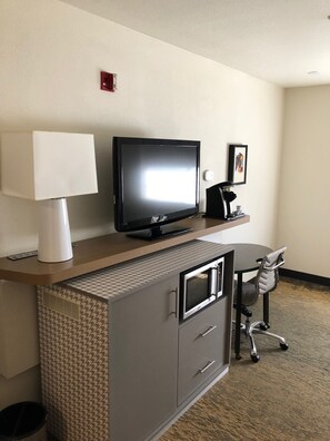 A hotel room with a flat-screen TV on a wooden console, a desk with a chair, and a microwave.