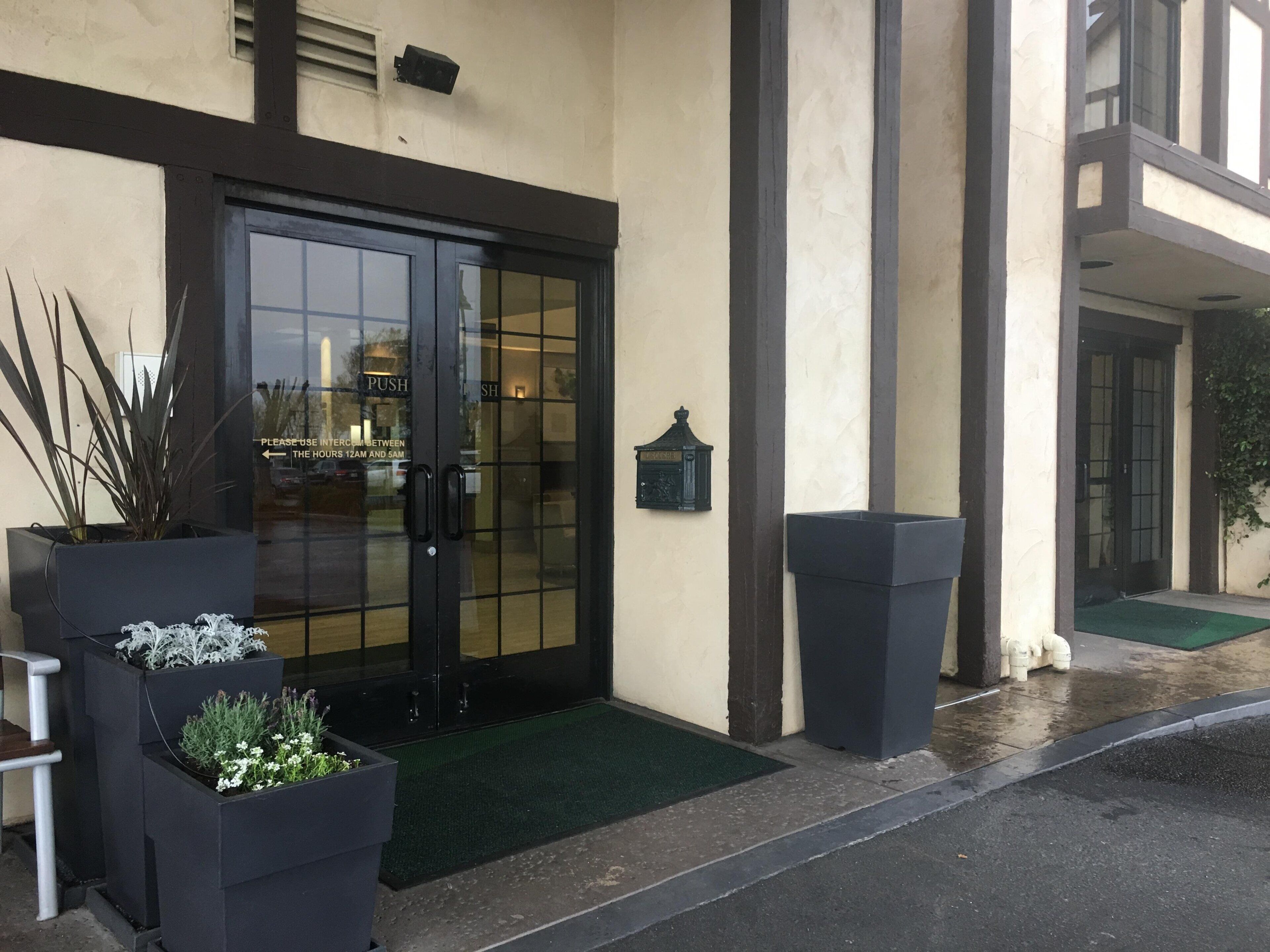 A building entrance with a glass door, potted plants, a small outdoor seating area, and a green mat.