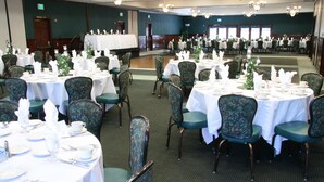 A banquet hall with round tables set for dining, green upholstered chairs, and chandeliers.