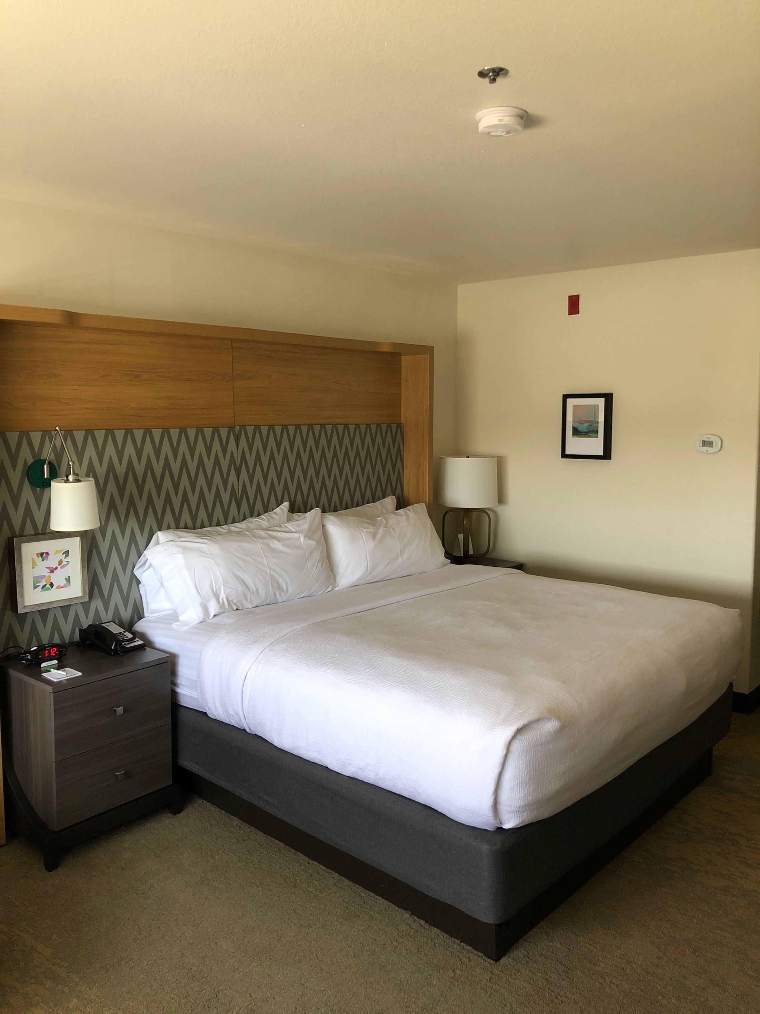 A hotel room with a bed, nightstands, a wall-mounted lamp, a framed picture, and a thermostat.