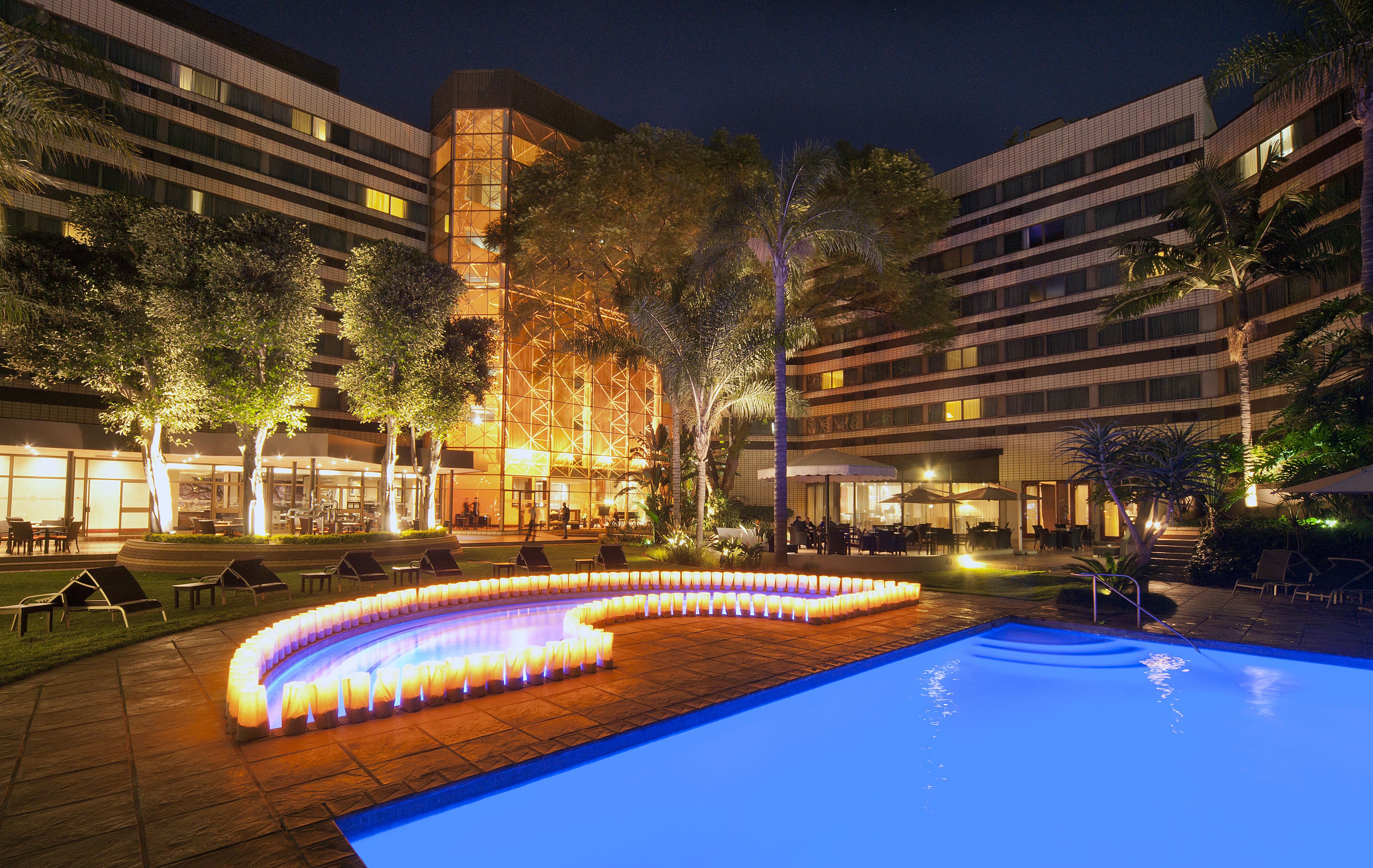 A nighttime pool area with a lit-up water feature, lounge chairs, and a building with multiple floors and balconies.