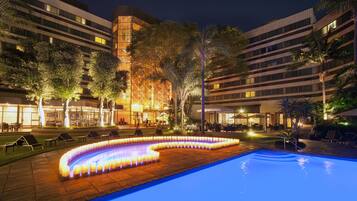 A nighttime pool area with a lit-up water feature, lounge chairs, and a building with multiple floors and balconies.