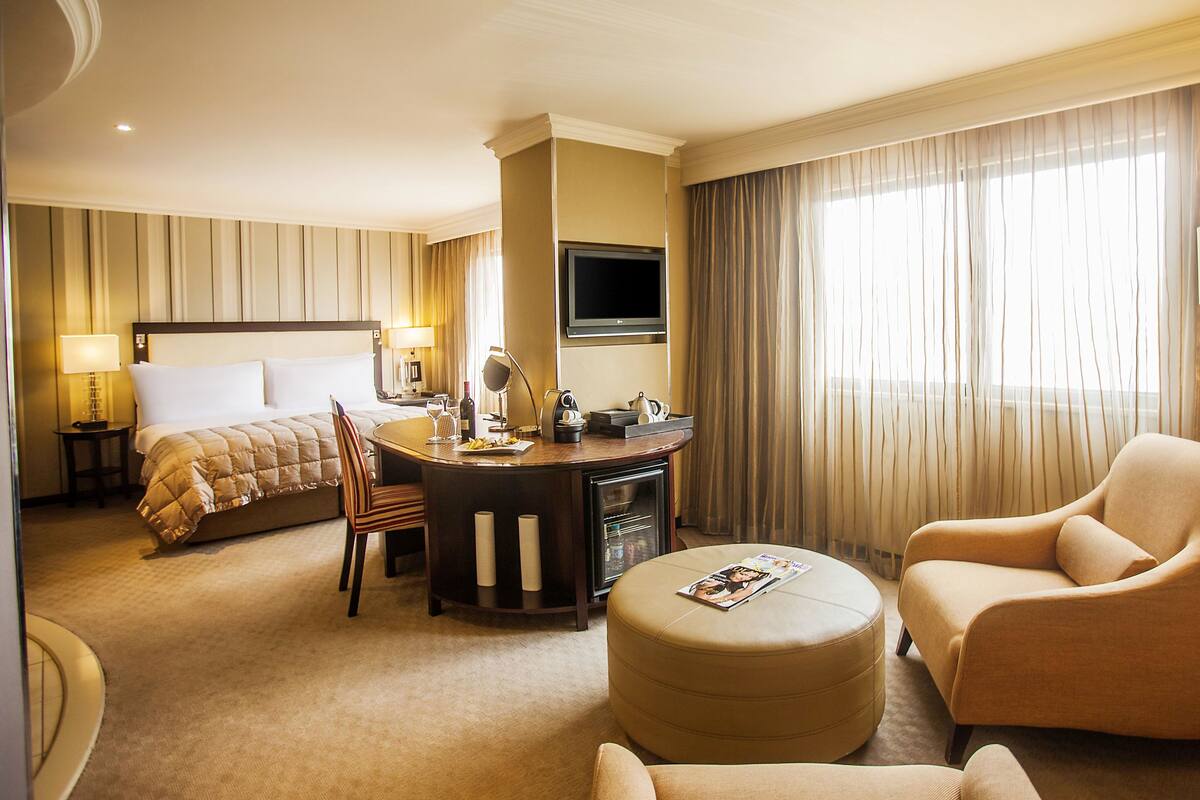 Habitación de hotel con cama, escritorio con silla, reposapiés redondo, sofá, televisor y lámpara.