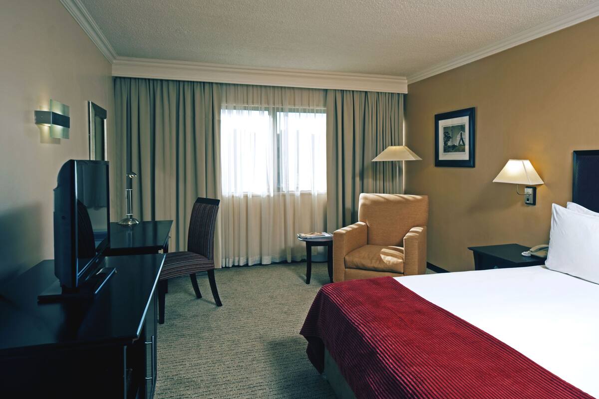 Habitación de hotel con cama, dos sillones, escritorio con computadora, televisor y ventana con cortinas.