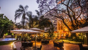An outdoor dining area with tables and chairs, surrounded by palm trees and a building with illuminated windows.