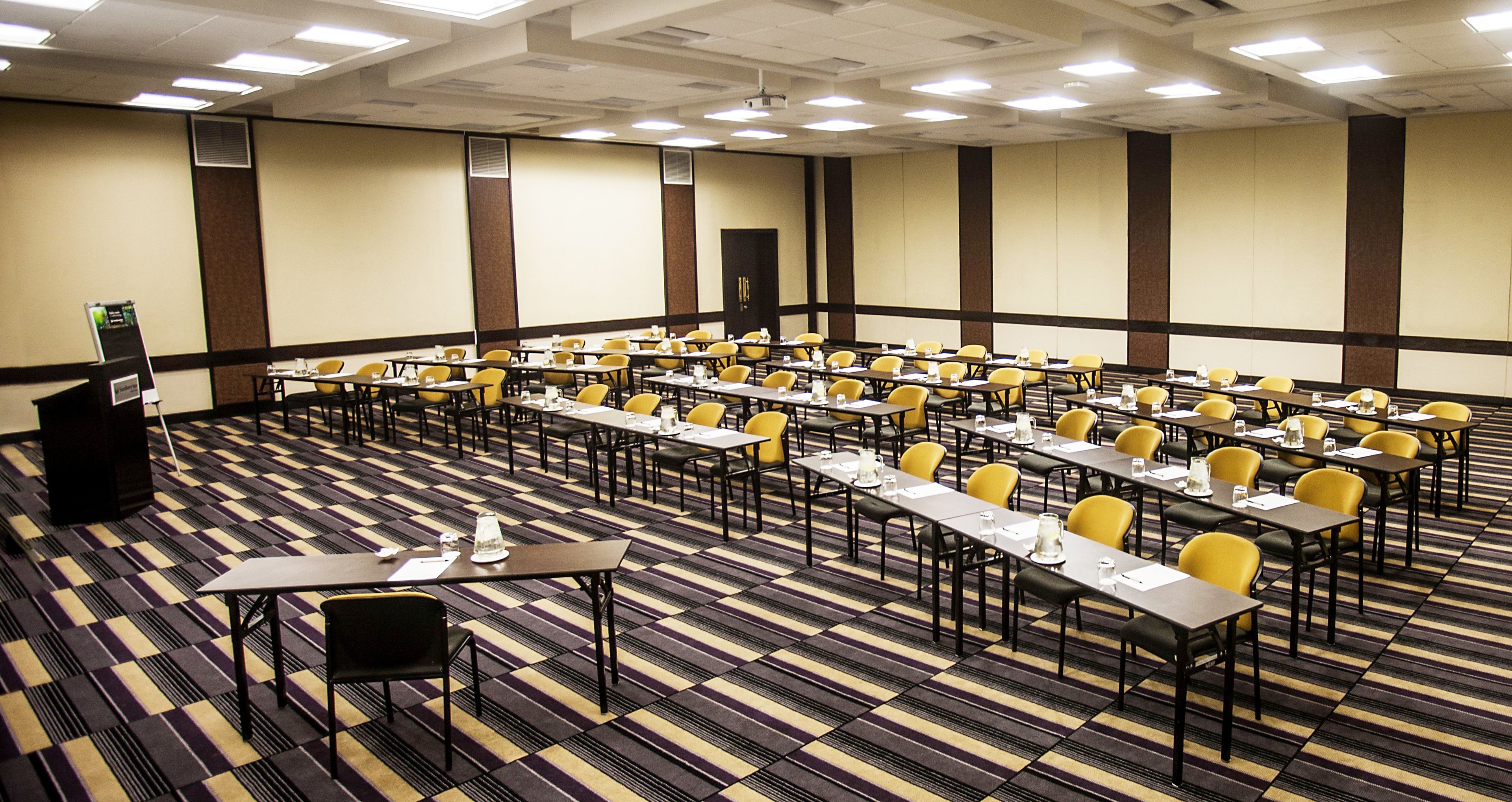 A conference room with rows of tables and chairs, a podium, and a carpeted floor.