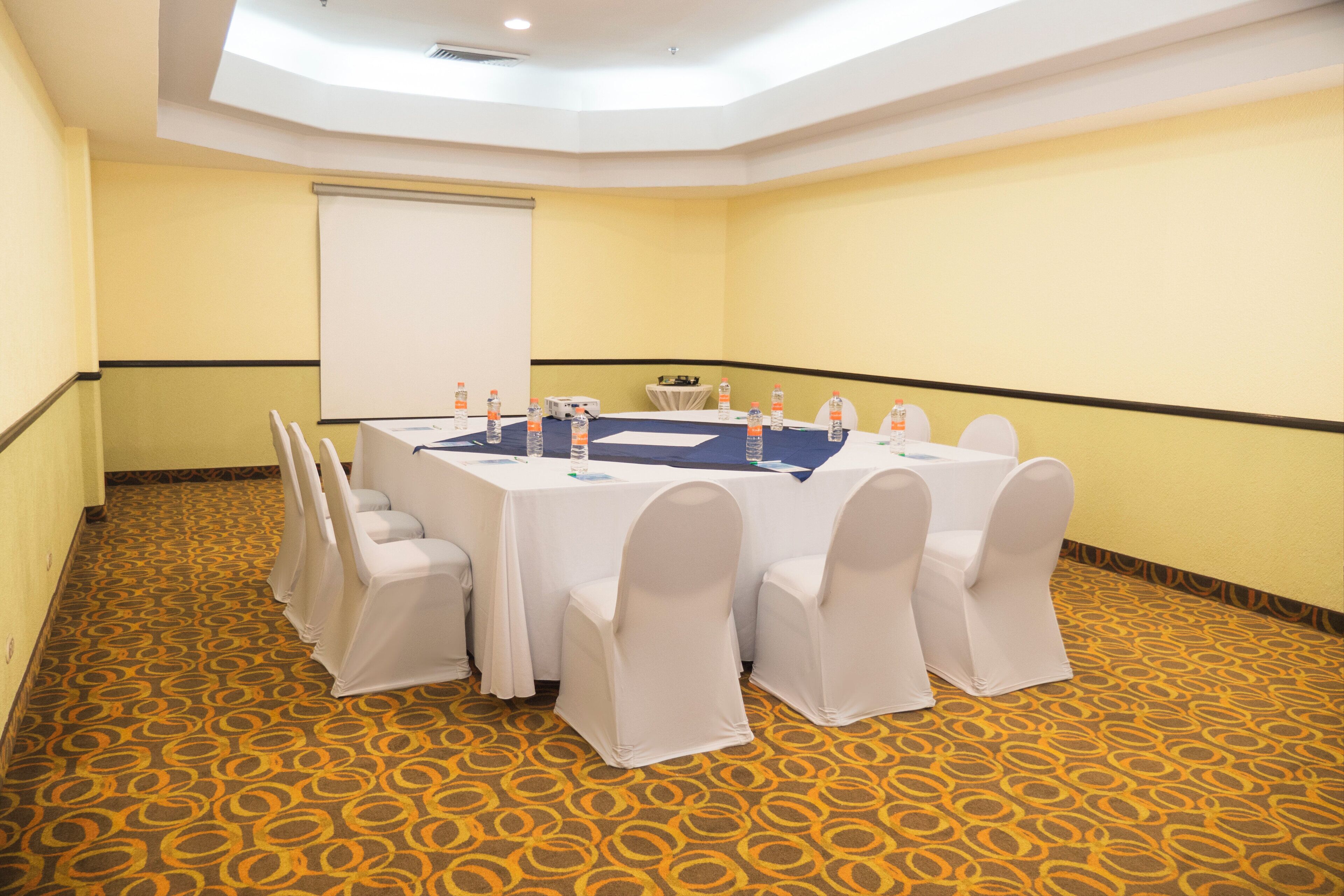 Ein Konferenzraum mit einem großen Tisch, weißen Stühlen und einer Projektionsleinwand.