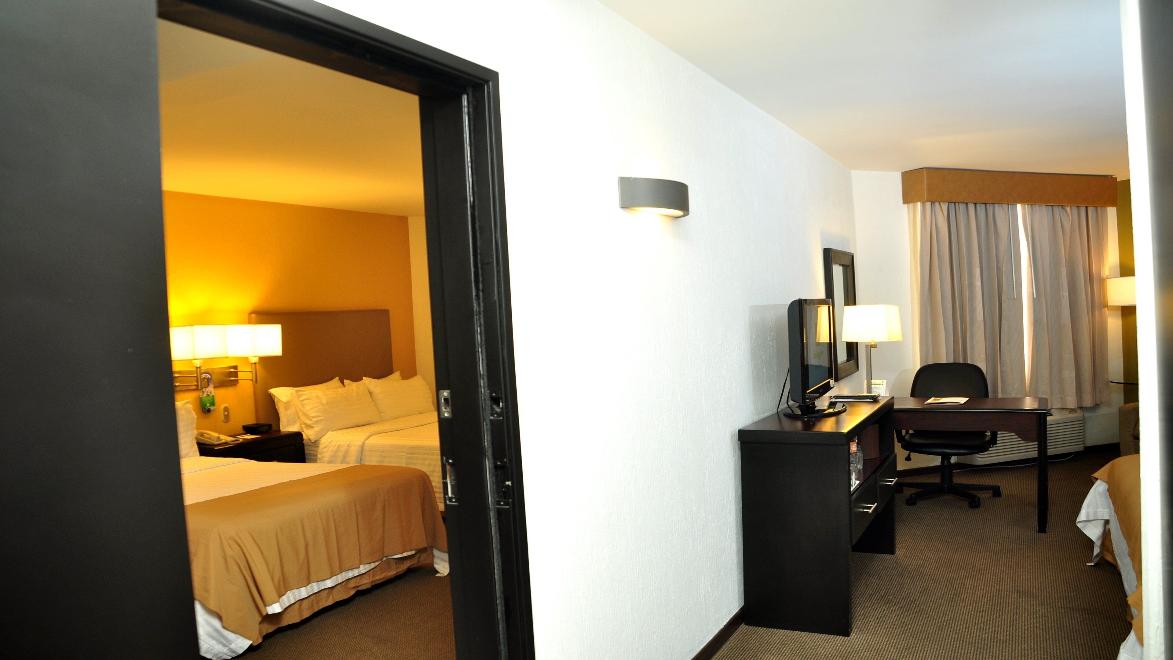Quarto de hotel com cama, escrivaninha, cadeira, luminária e janela com cortinas.