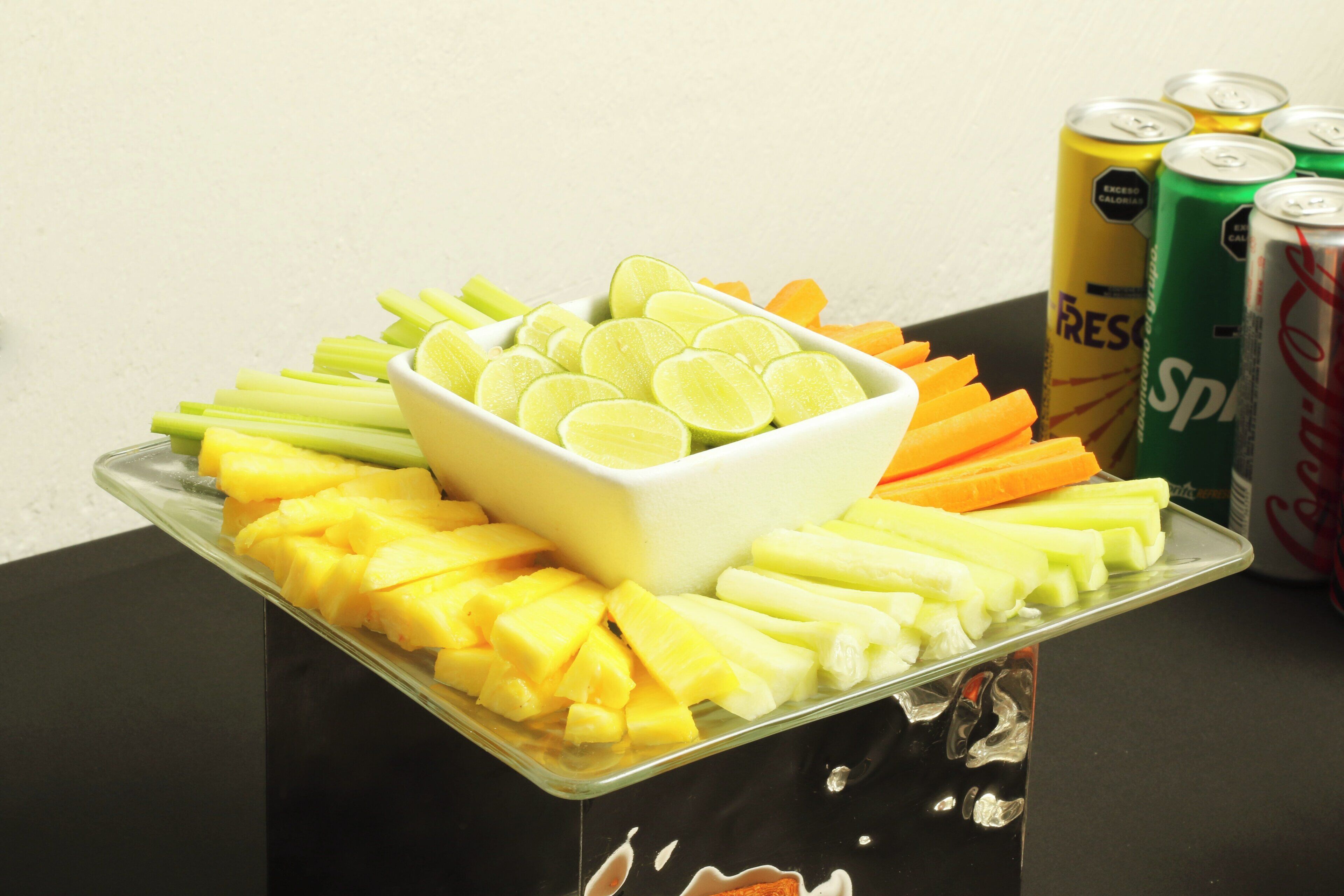 Un plateau contenant un assortiment de fruits et de légumes est posé sur une table, avec des canettes de soda en arrière-plan.