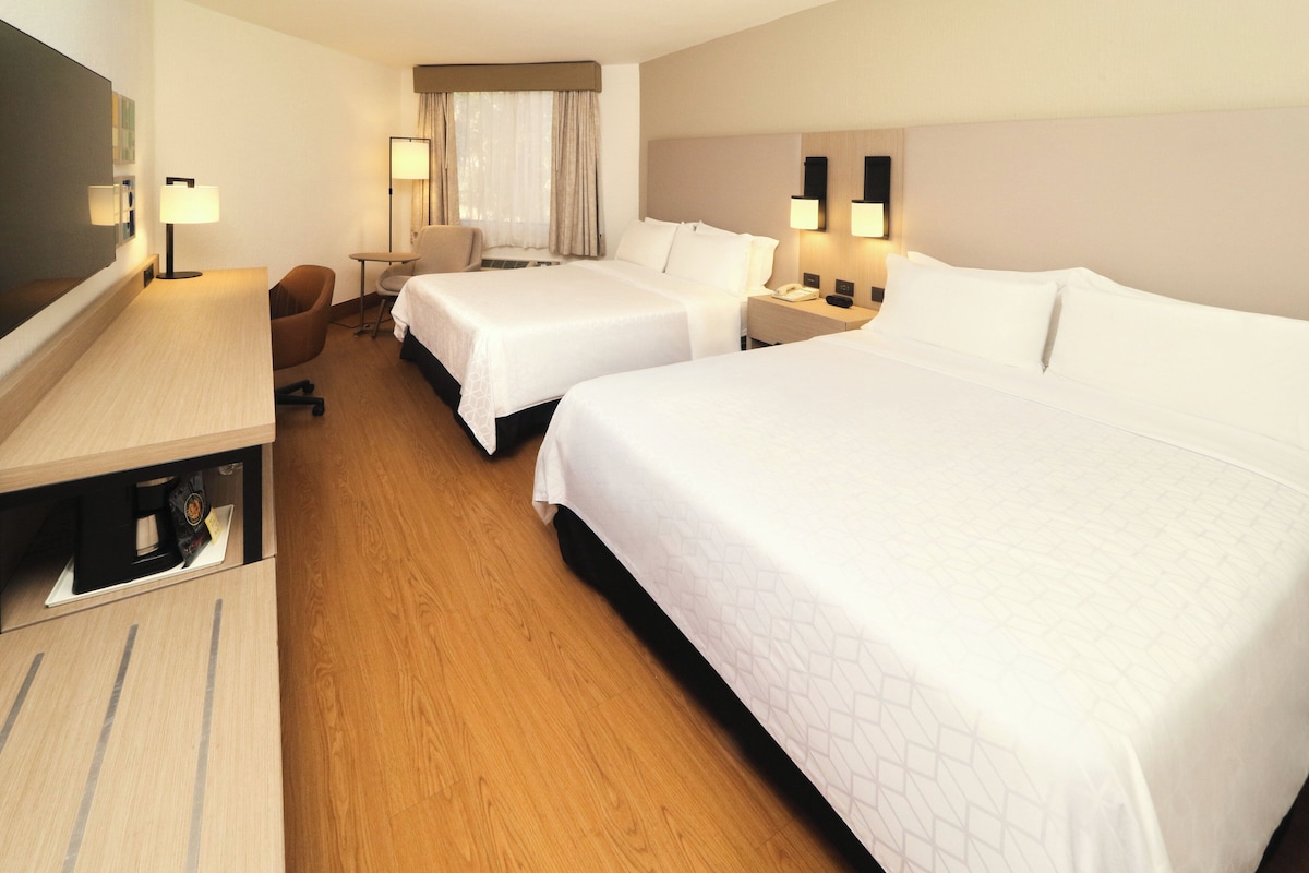 A hotel room with two beds, a desk, a chair, a desk lamp, a nightstand, and a window with curtains.