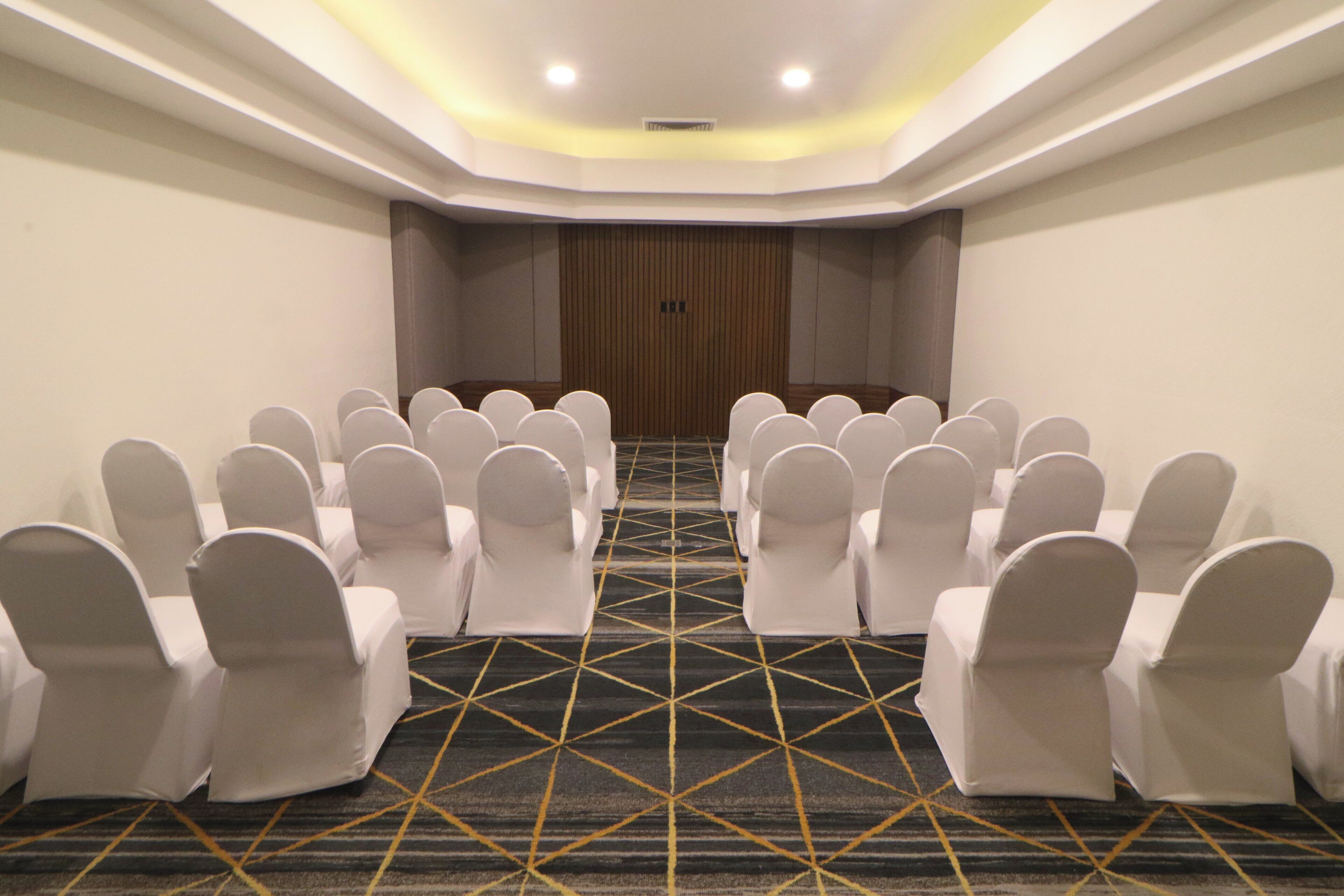 Uma sala de conferências com fileiras de cadeiras brancas, um tapete estampado e uma parede de painéis de madeira.