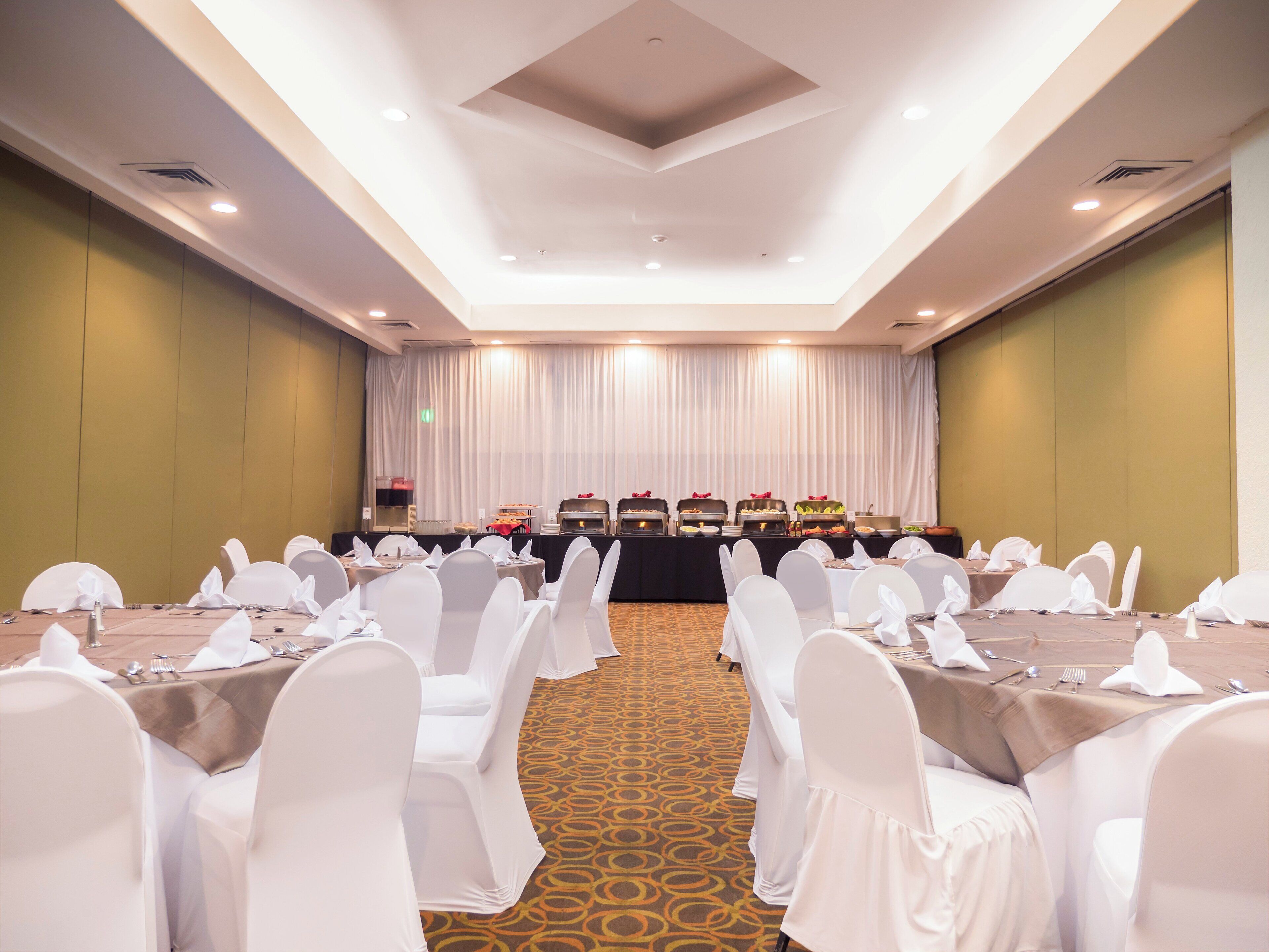 Un salón de conferencias con mesas y sillas dispuestas en filas, una mesa de buffet con diferentes alimentos, y un escenario con cortinas.