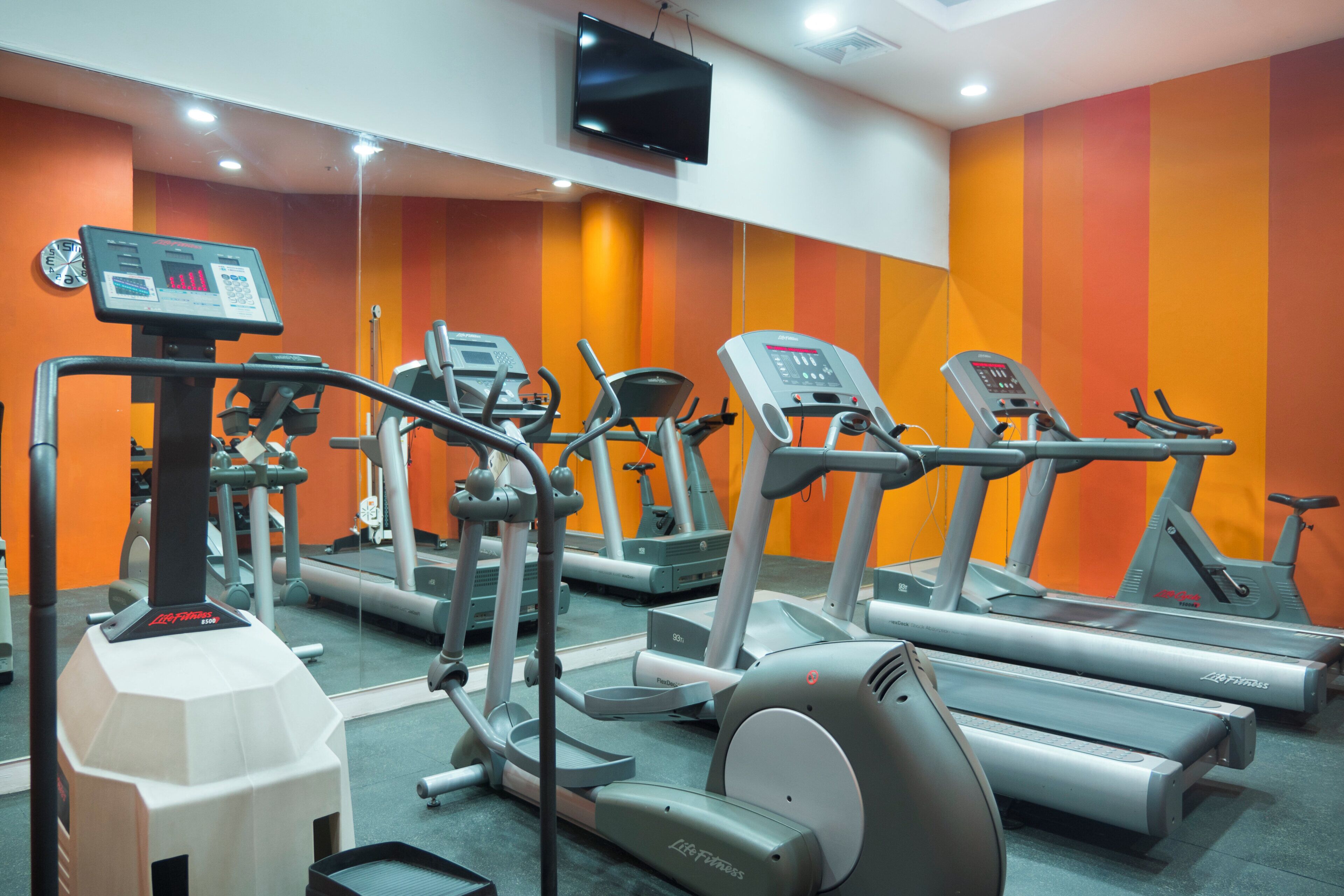 A gym with various cardio machines, a digital display, and a television mounted on the wall.