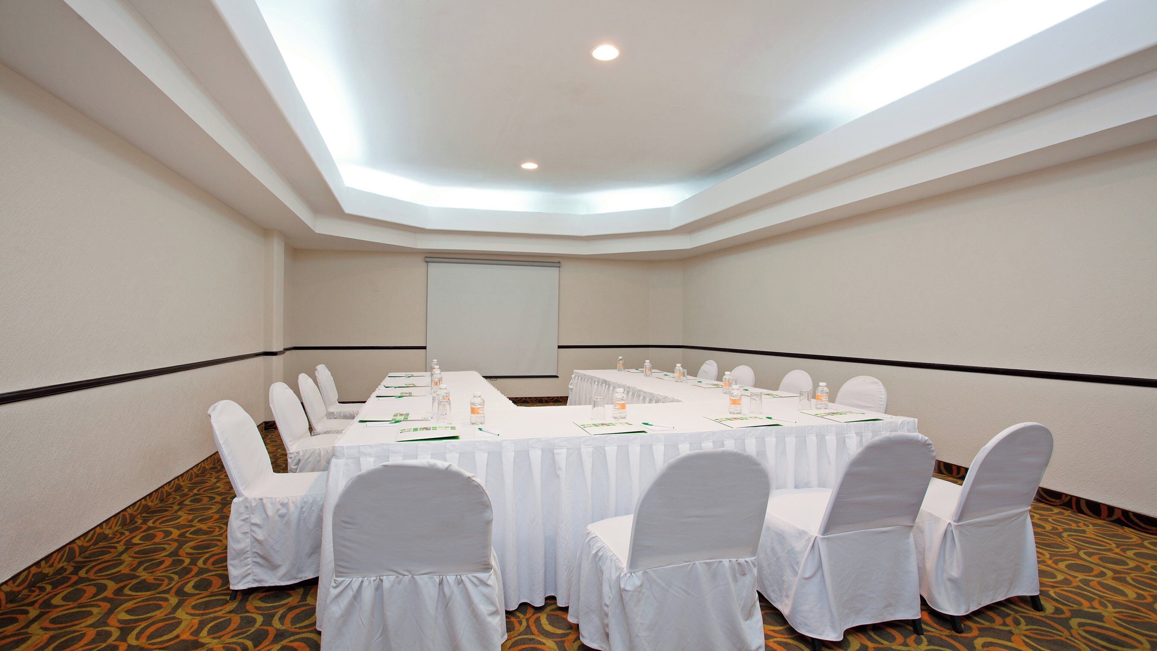 Une salle de conférence avec une longue table, des chaises blanches et un écran de projection.