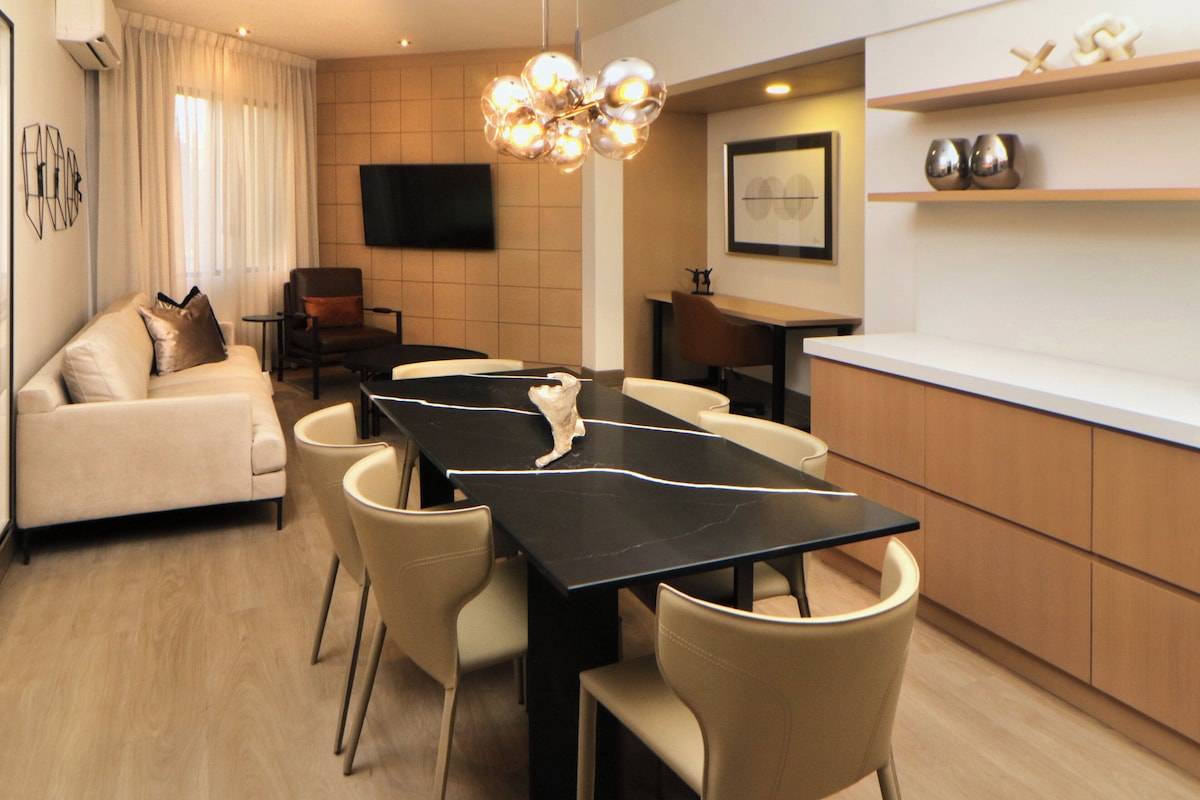 A modern living room with a sofa, a dining table with chairs, a kitchen area with a countertop, and a flat-screen TV mounted on the wall.