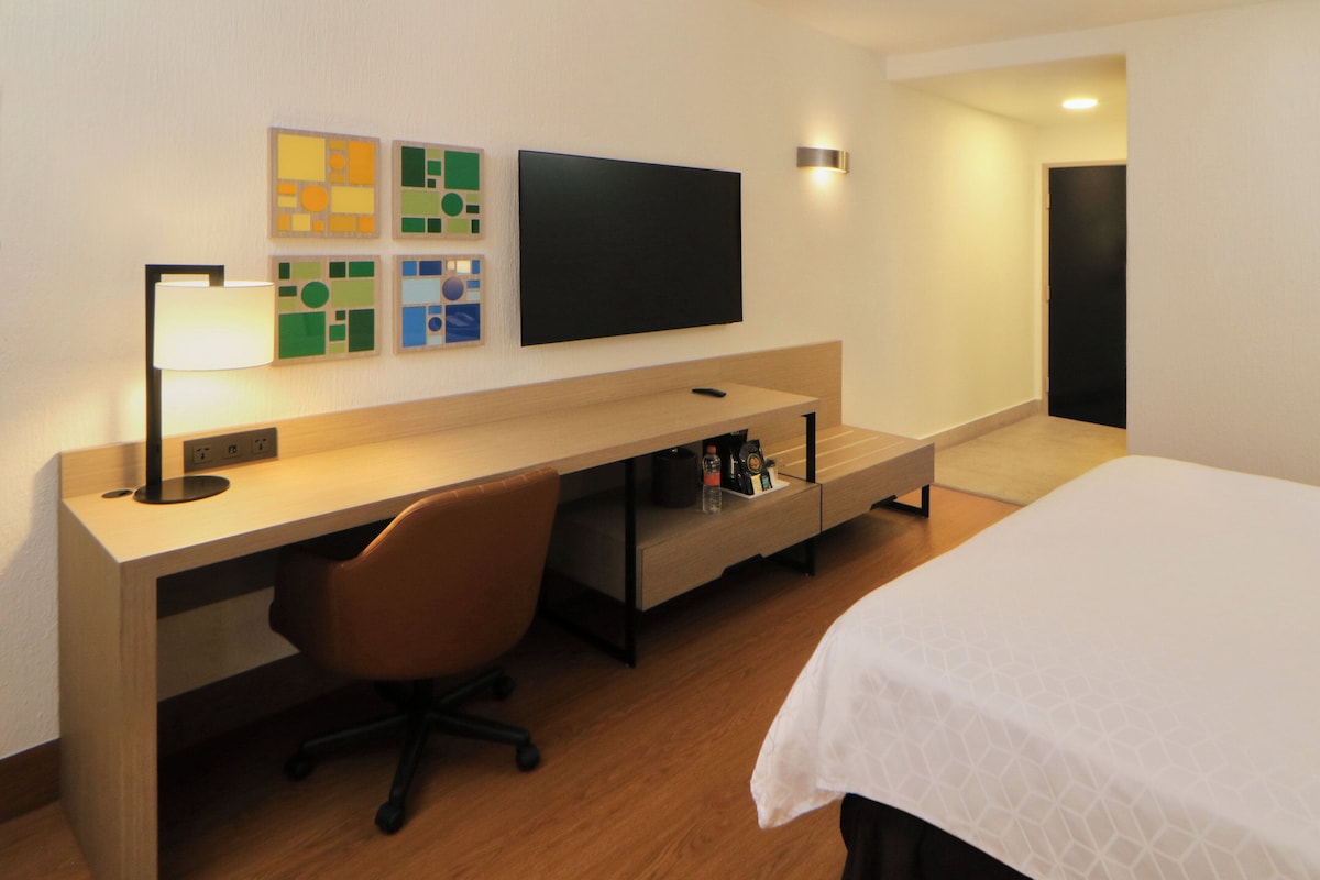 A hotel room with a bed, a desk with a chair, a flat-screen TV, and artwork on the wall.