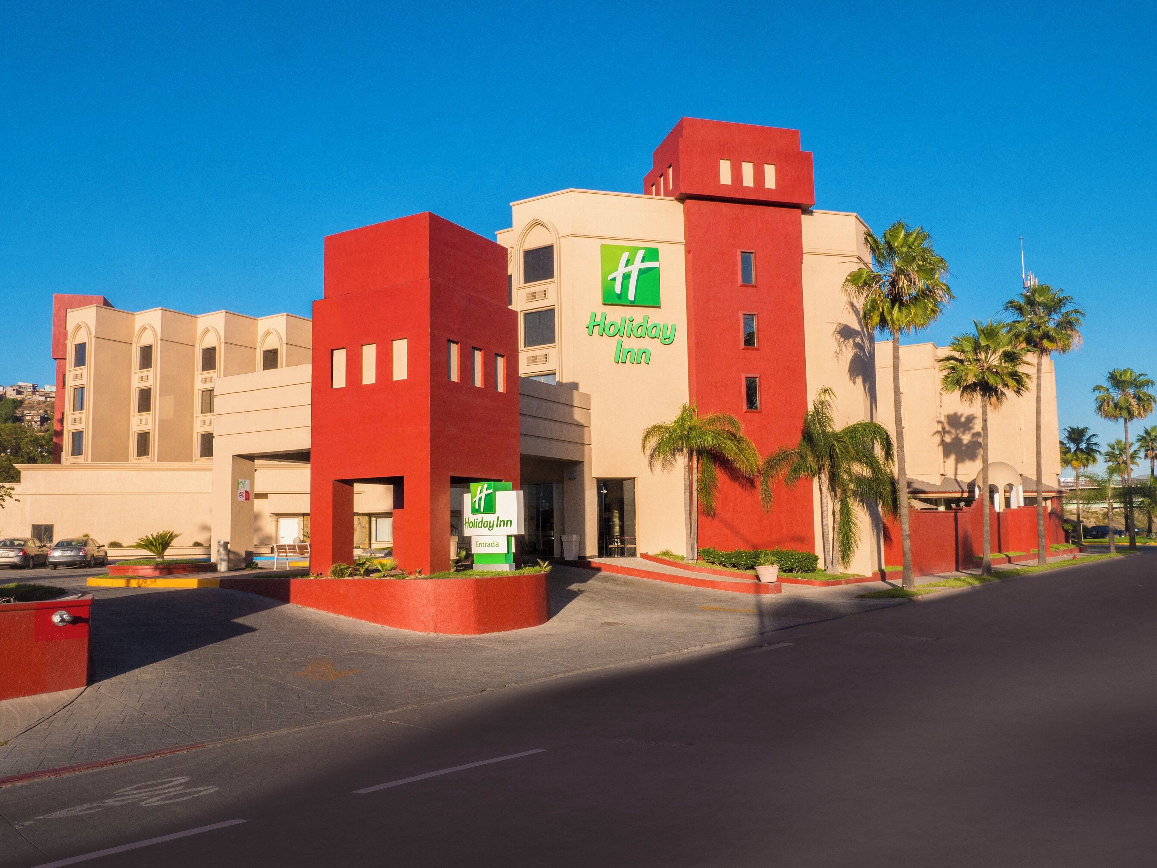 Un hotel Holiday Inn con fachada roja y color beige, palmeras y un cielo azul despejado.