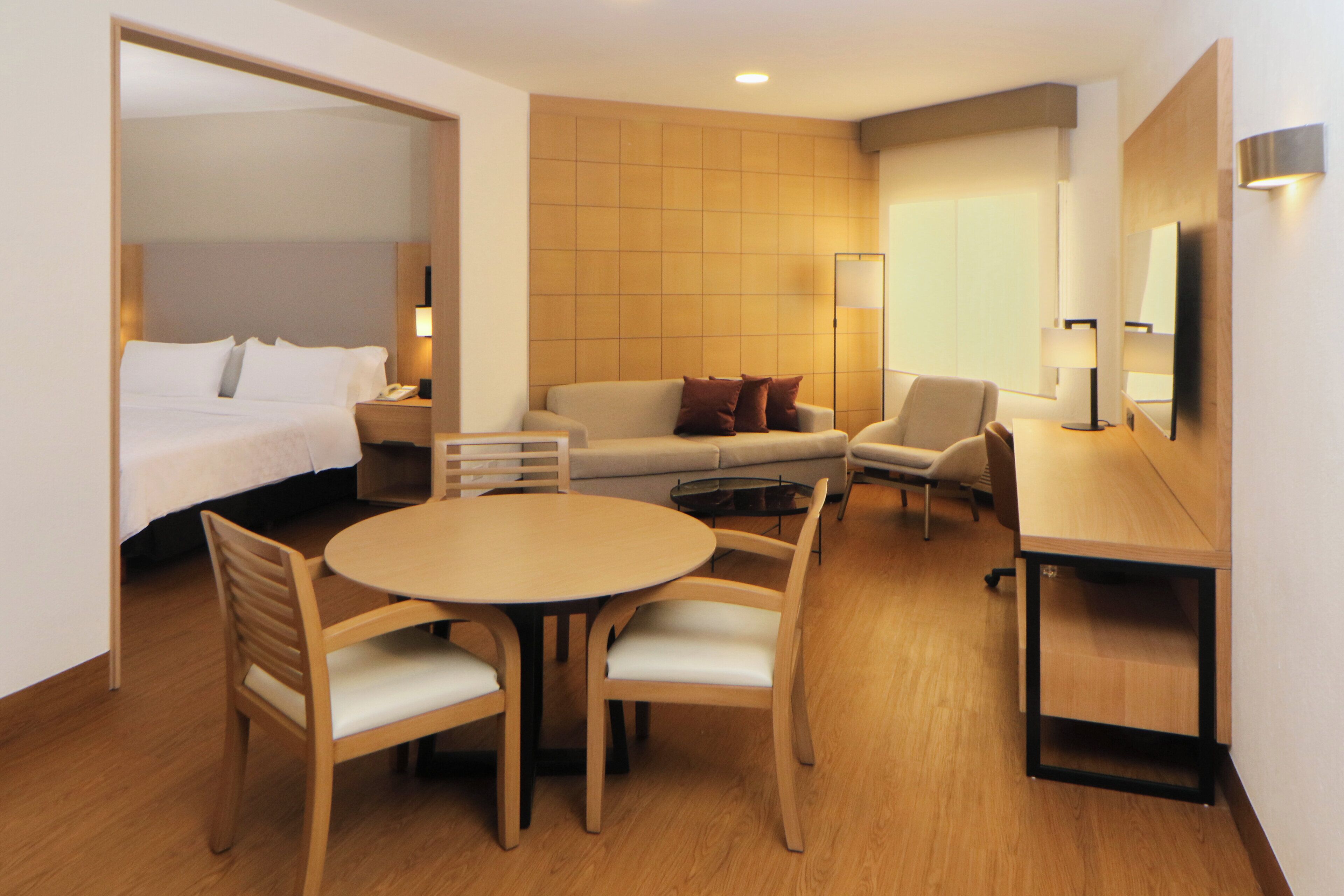 A hotel room with a bed, a sofa, a dining table with chairs, a TV, and a lamp.