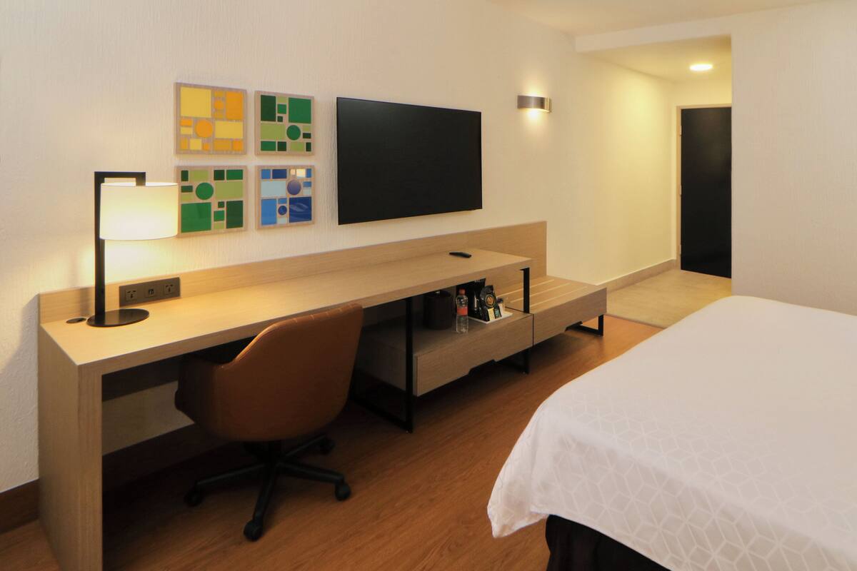 Een hotelkamer met een houten bureau, een stoel, een bed met een witte spreidek en een wandtelevisie, plus een lamp bevestigd aan de muur.