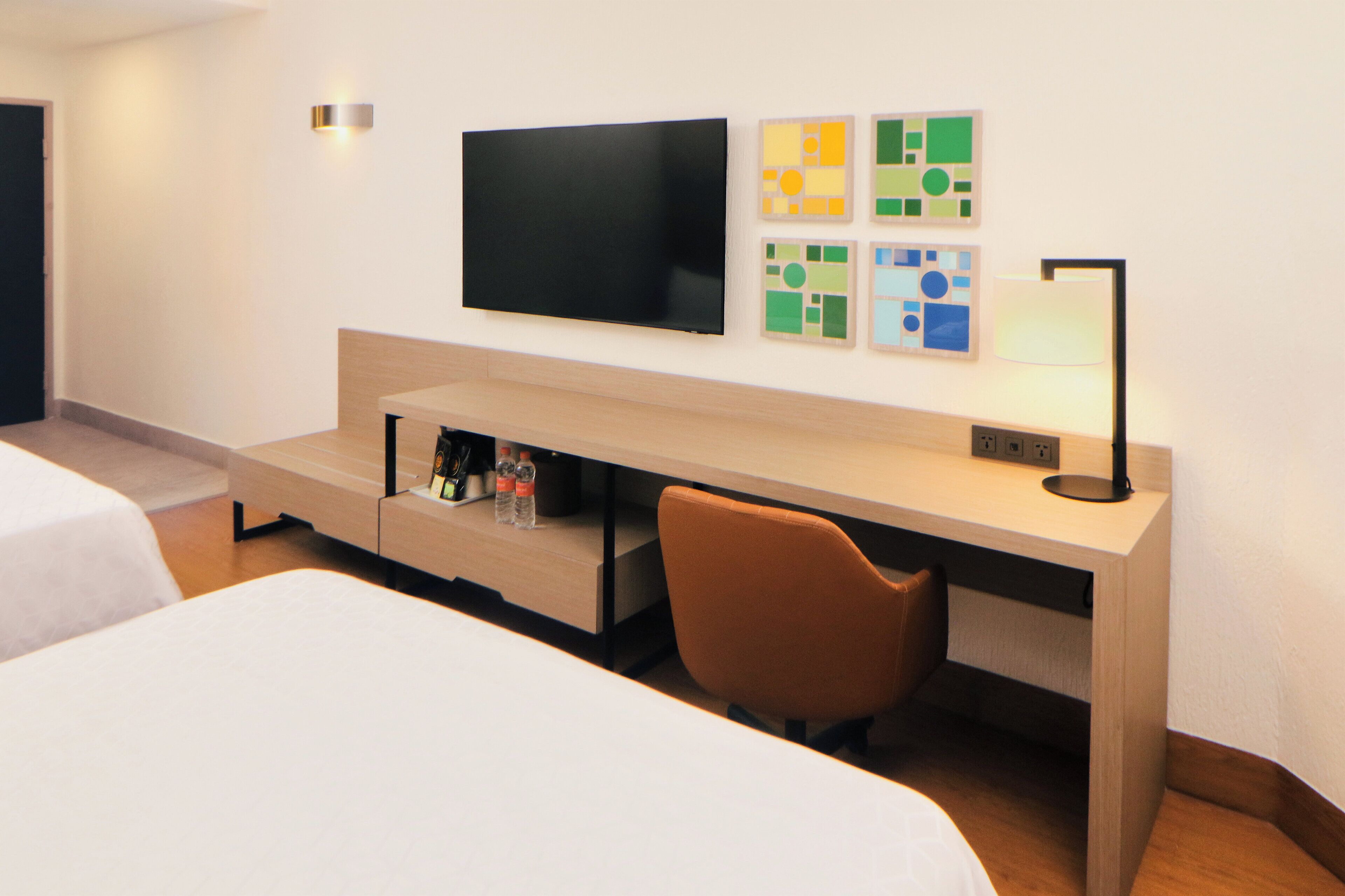 A hotel room with a bed, a desk with a chair, a flat-screen TV, and artwork on the wall.