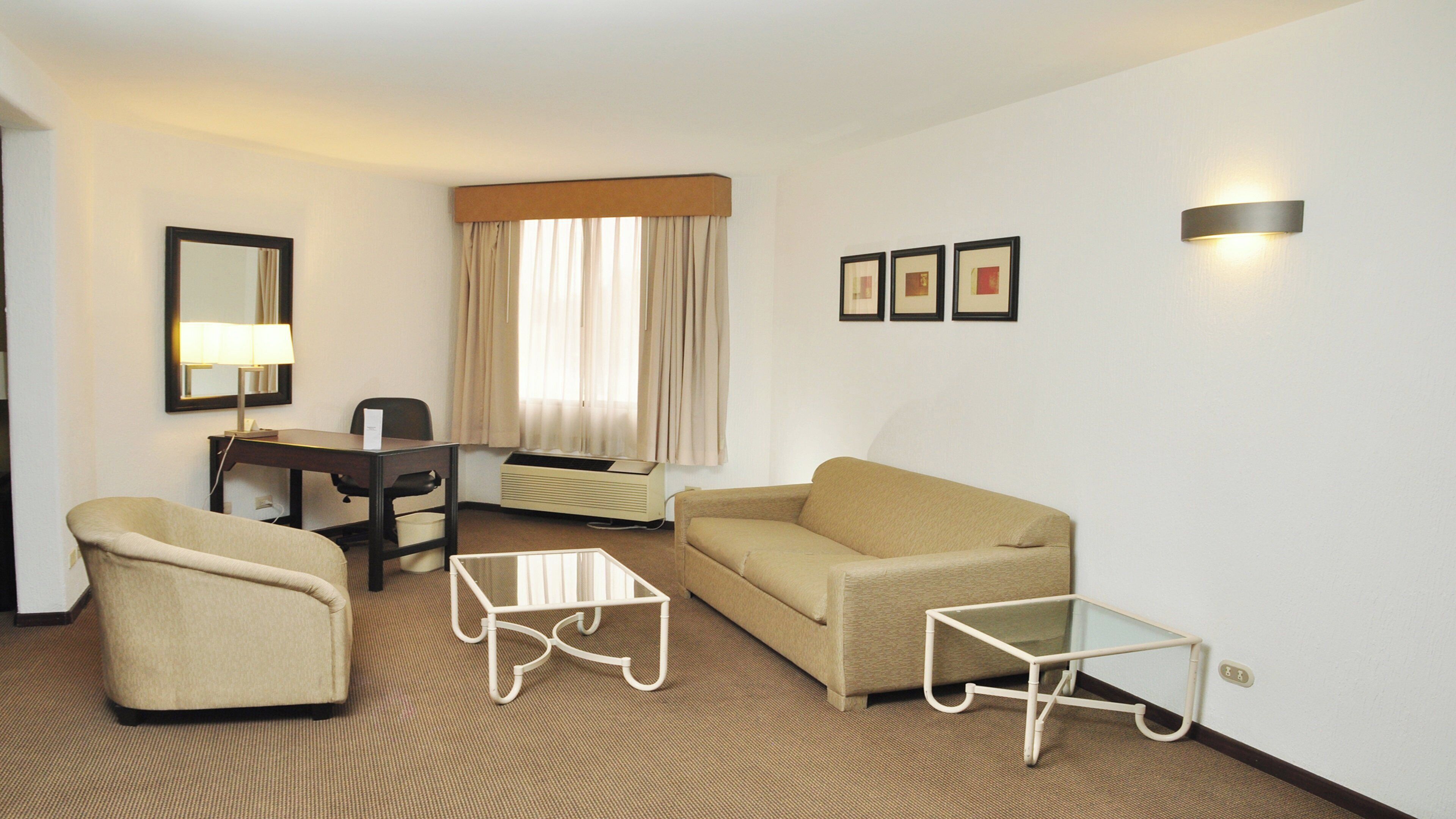 A hotel room with a sofa, armchair, small table, and a desk with a lamp.