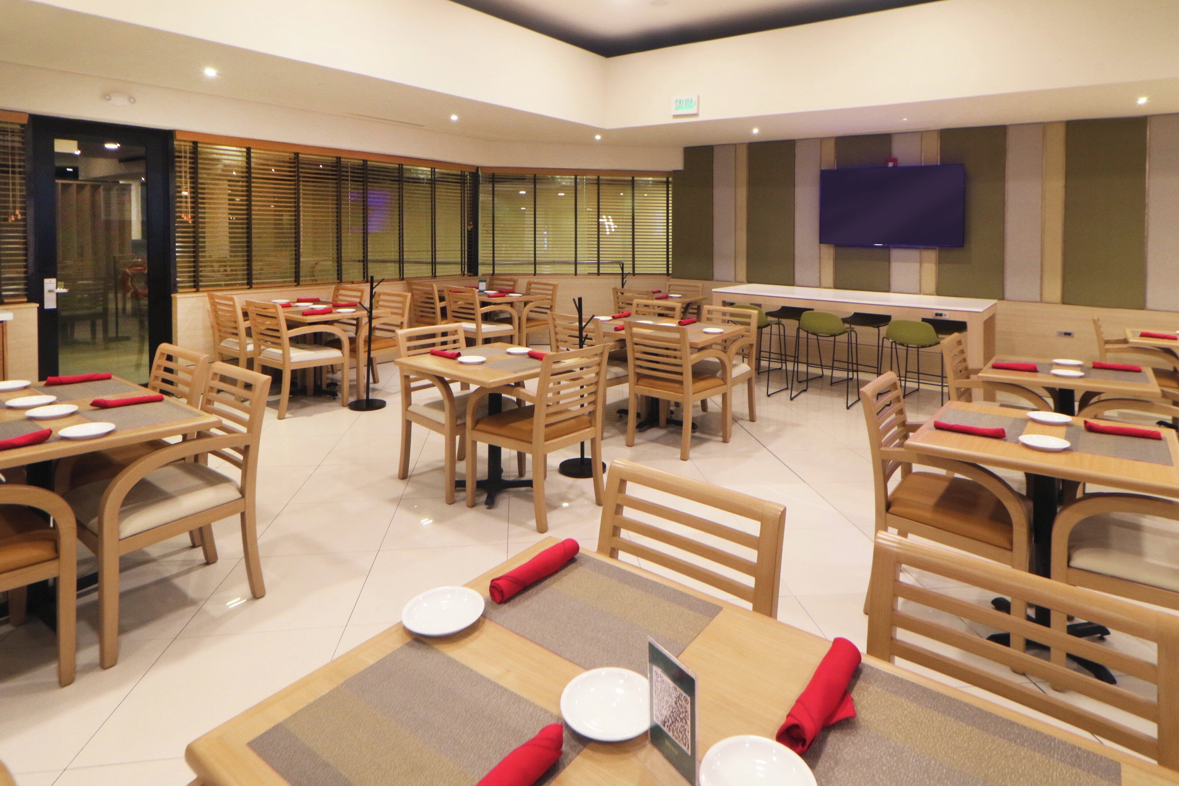A spacious dining area with multiple tables and chairs, a flat-screen TV mounted on the wall, and a large window with blinds.