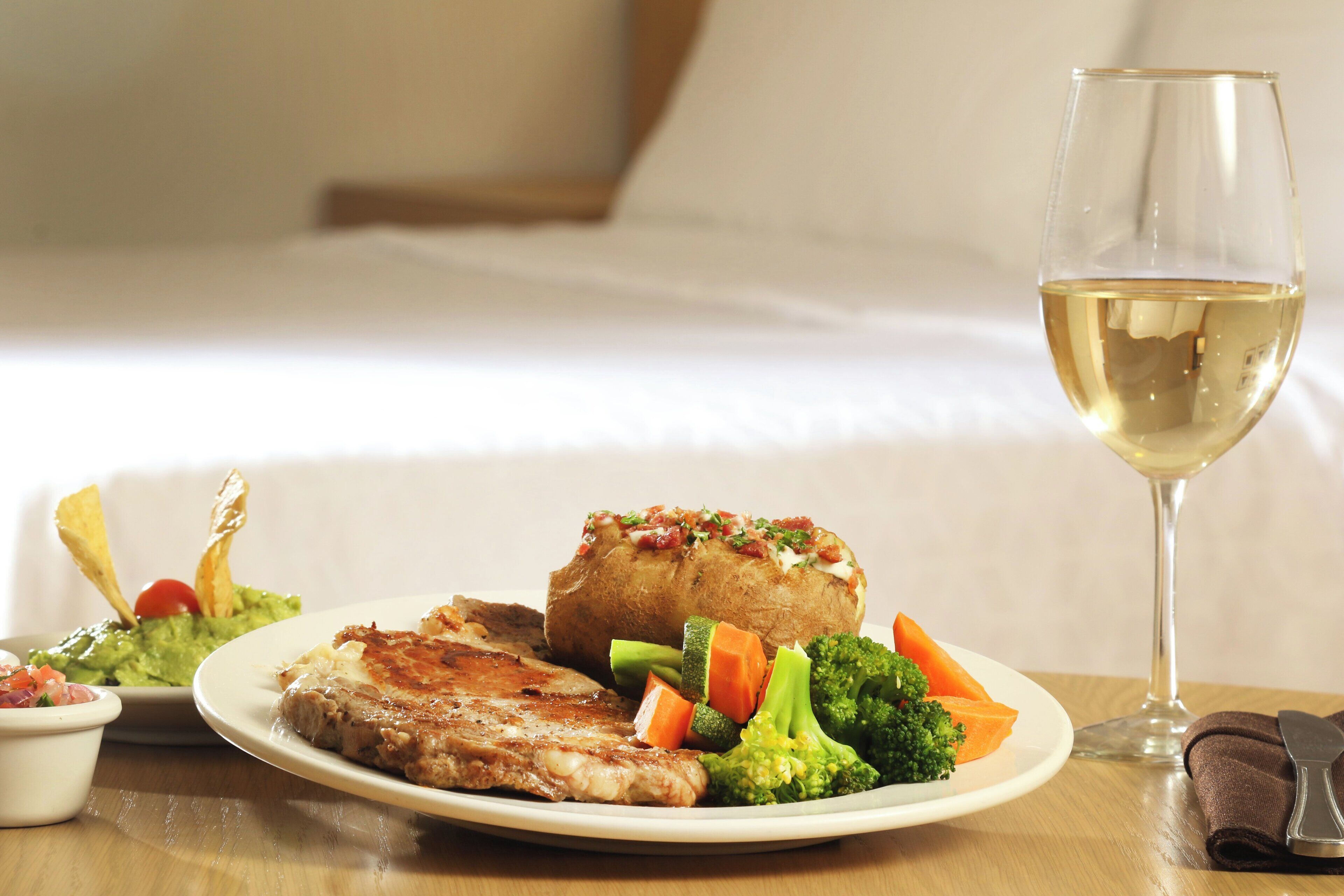 A plate of food including steak, vegetables, and a baked potato, with a glass of white wine on a table.