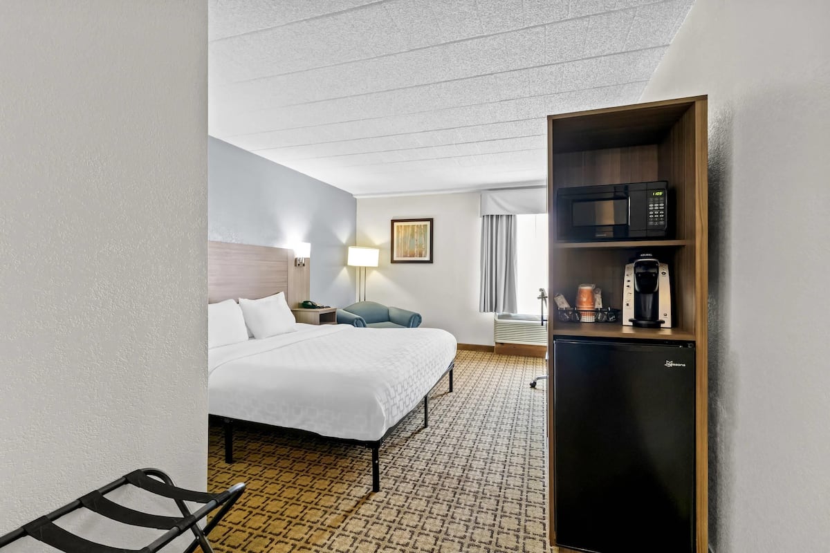 A hotel room with a bed, a desk, a chair, a microwave, a coffee maker, and a refrigerator.