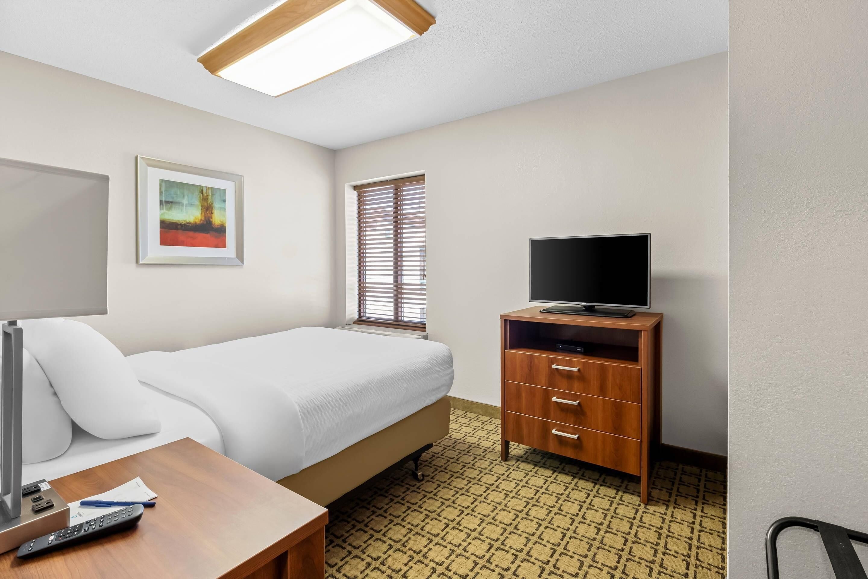 Quarto de hotel com cama, televisão sobre suporte de madeira, escrivaninha com luminária, quadro na parede e janela com persianas.