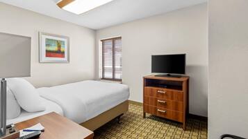 Quarto de hotel com cama, televisão sobre suporte de madeira, escrivaninha com luminária, quadro na parede e janela com persianas.