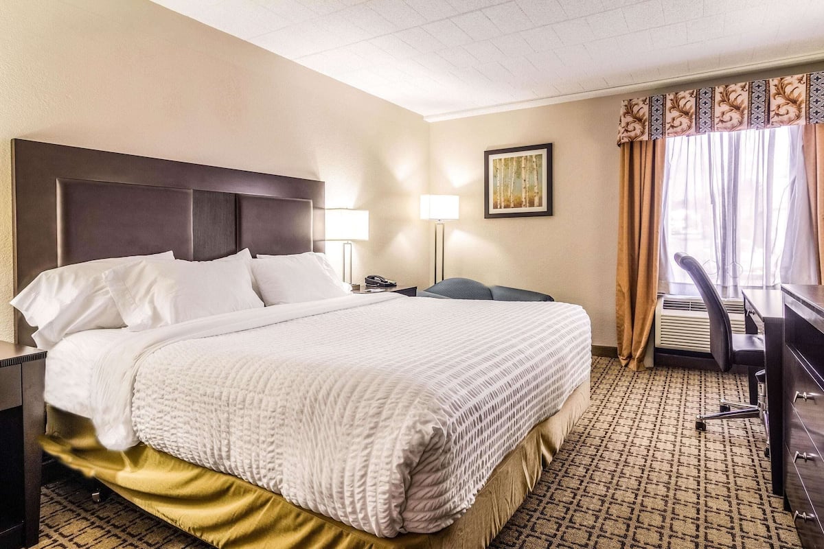 A hotel room with a large bed, bedside lamps, a desk, and an air conditioning unit.