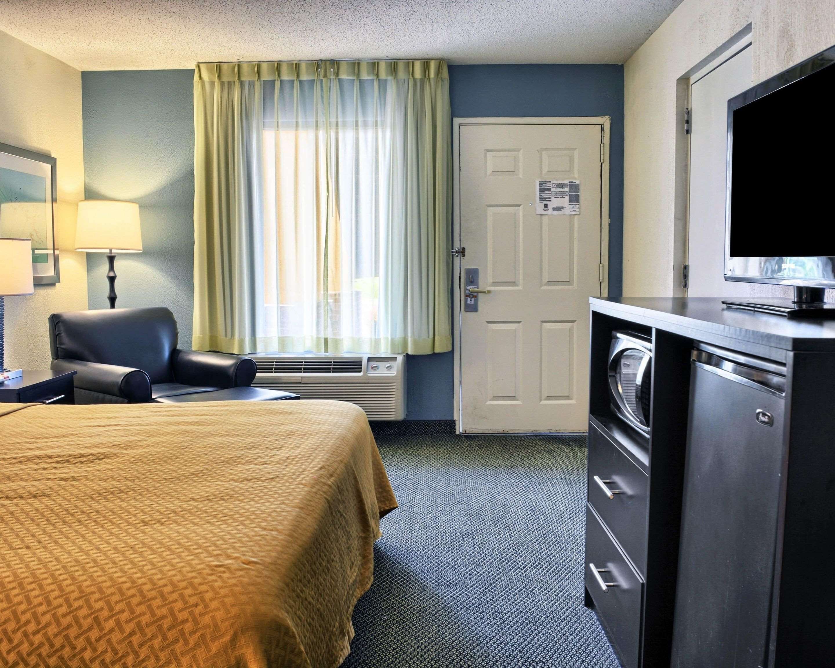 A hotel room with a bed, a chair, a desk with a lamp, a television, and a washer and dryer.