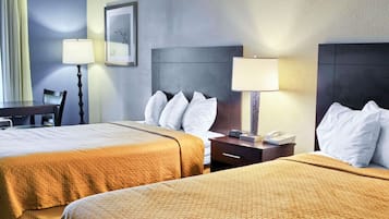 A hotel room with two beds, a desk, a chair, and a nightstand with a lamp.