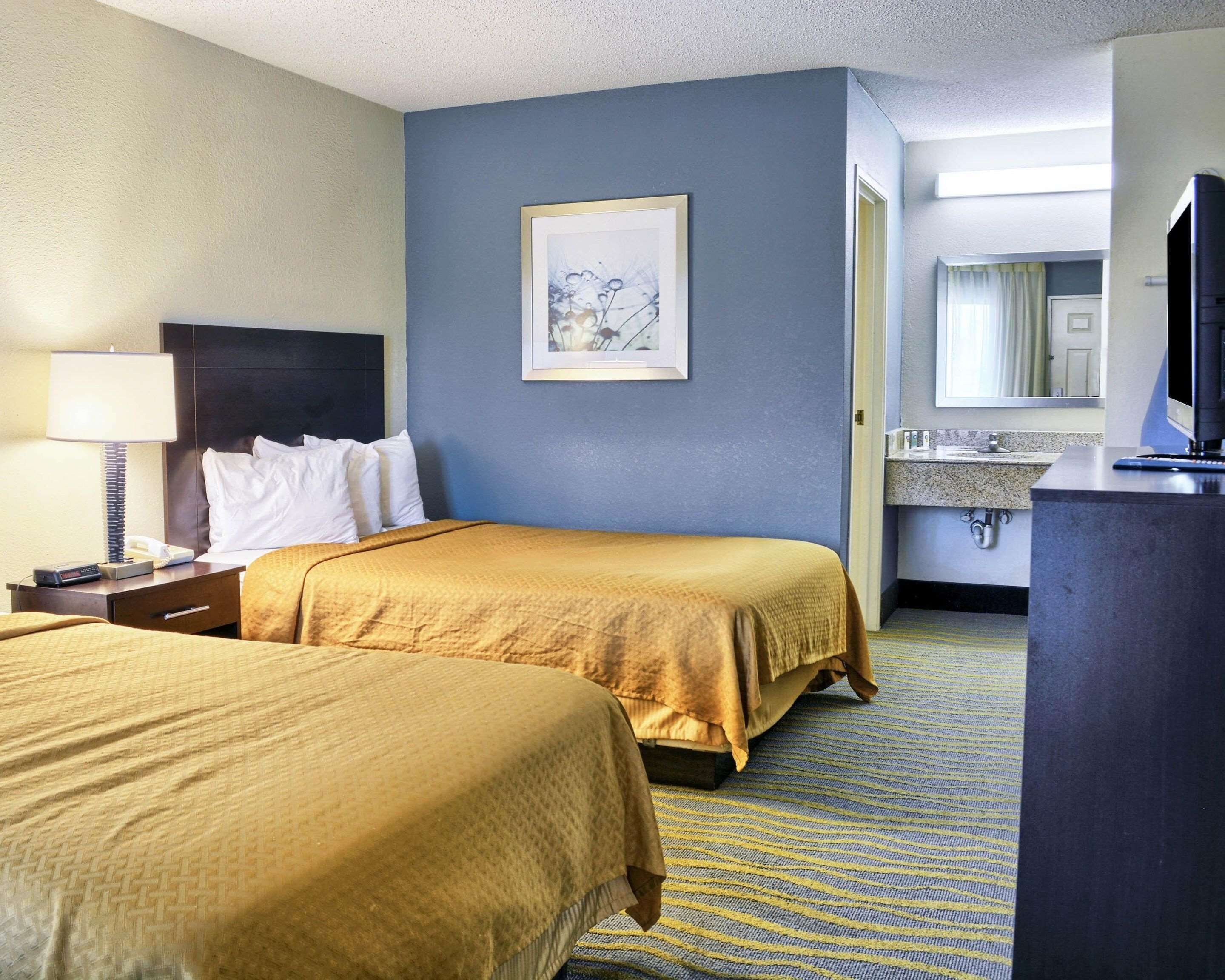 A hotel room with two beds, a nightstand with a lamp, a framed picture on the wall, and a television on a stand.