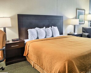 A hotel room with a large bed, a dark headboard, white pillows, a brown blanket, a desk with a lamp, and a chair. - Quality Inn Thornburg (Woodford)
