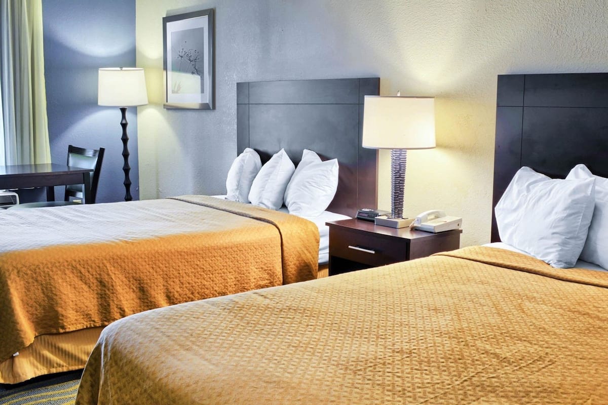 A hotel room with two beds, a desk, a chair, a lamp, and a framed picture on the wall.