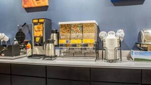 A breakfast bar with waffle maker, coffee maker, cereal dispensers, and dishware. - Econo Lodge Ankeny - Des Moines (Ankeny)