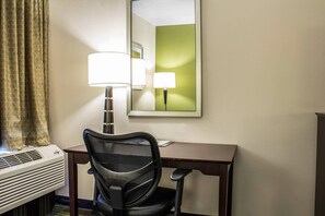 A hotel room with a desk, chair, air conditioning unit, lamp, and a mirror reflecting a green wall. - Econo Lodge Ankeny - Des Moines (Ankeny)