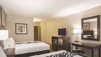 Habitación de hotel con dos camas, un escritorio con televisor, un espejo y una puerta que da a otra habitación.