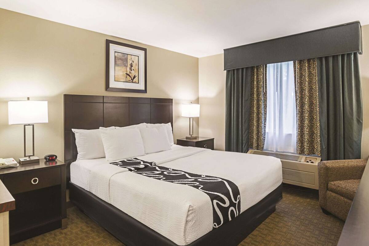 Een hotelkamer met een groot bed, twee nachtkastjes, een stoel, een raam met gordijnen en een ingelijste foto aan de muur.