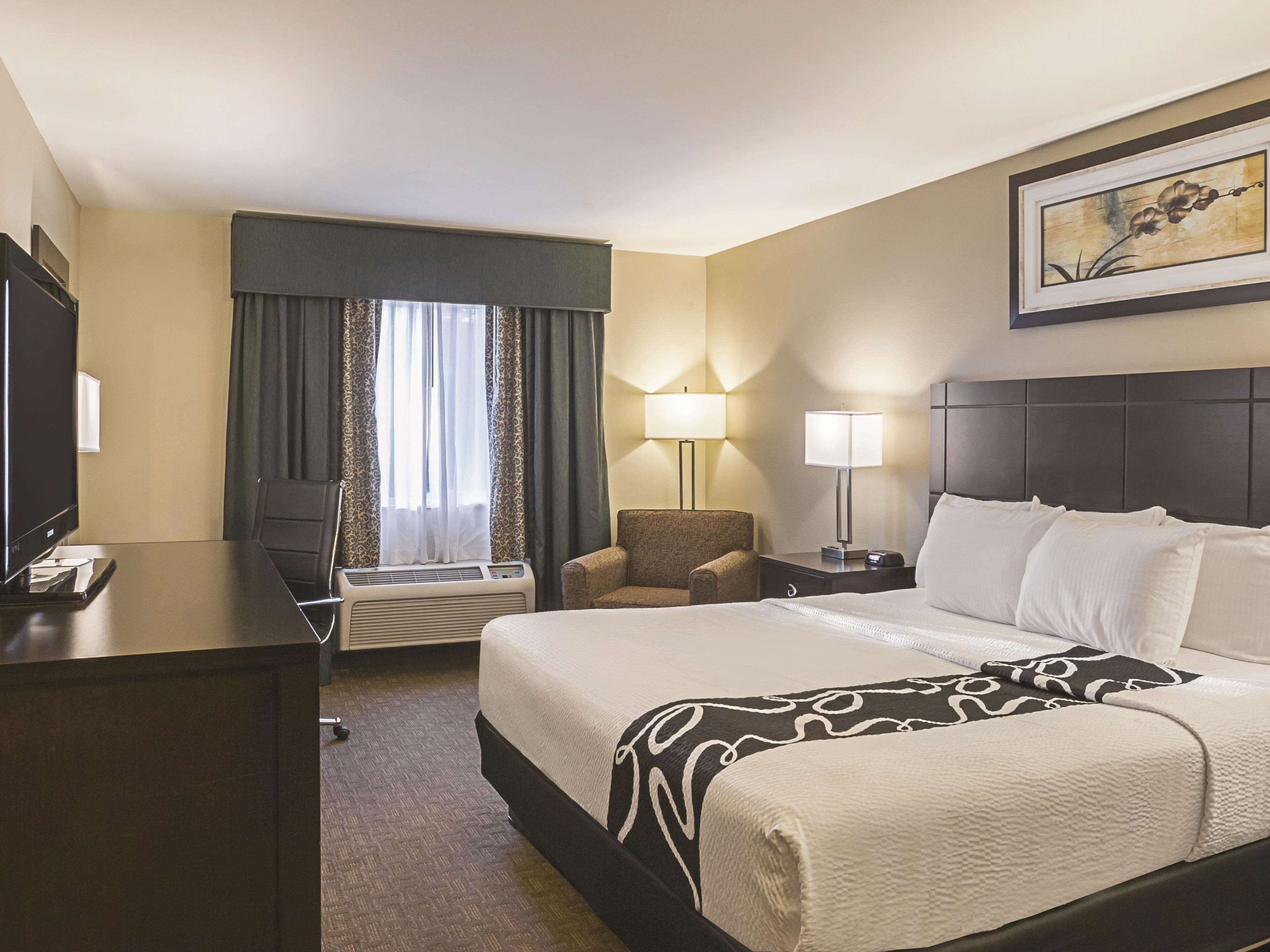Habitación de hotel con una cama grande, dos mesitas de noche con lámparas, un escritorio con televisor, una silla y una ventana con cortinas.