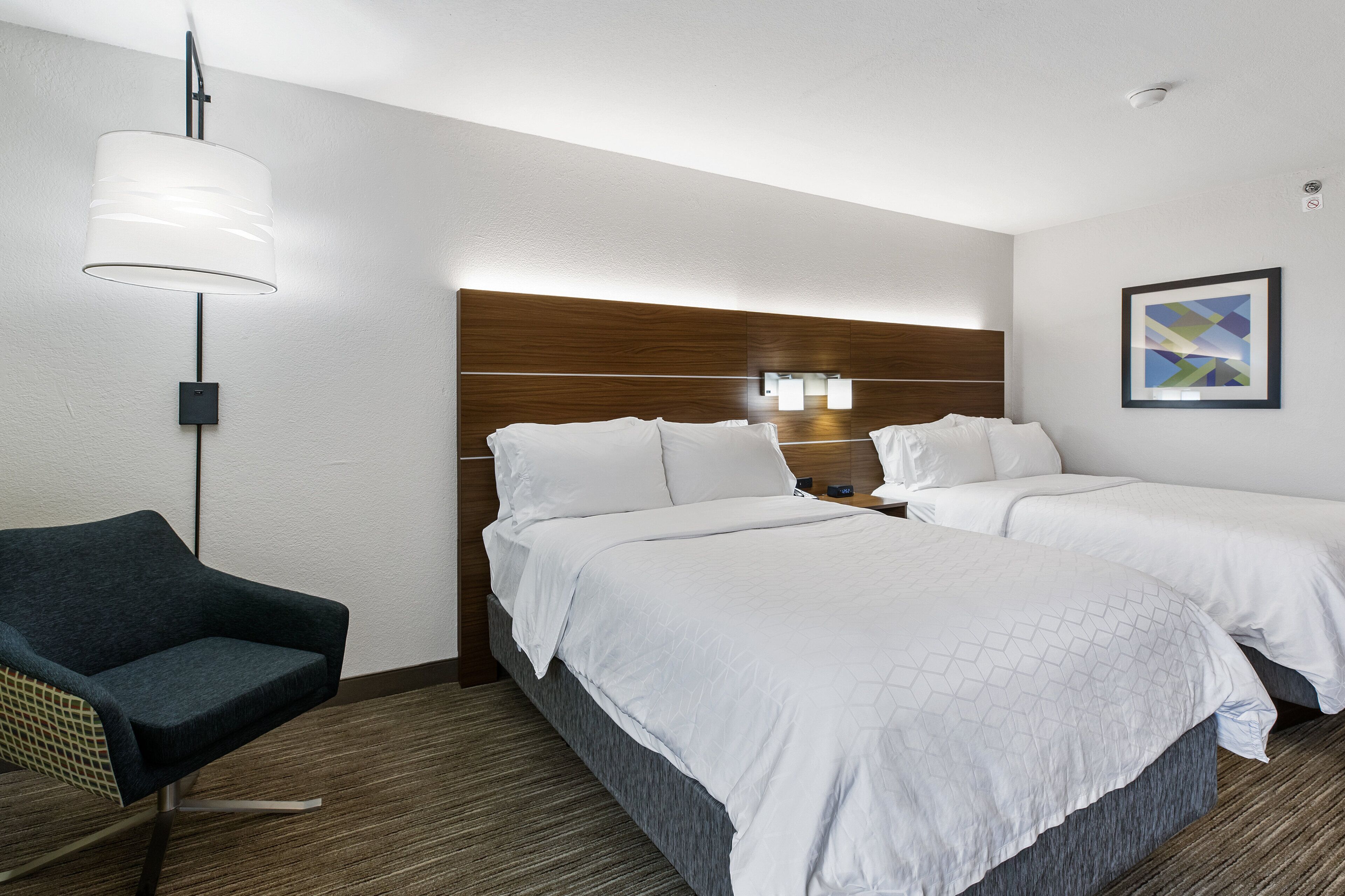 Une chambre d’hôtel avec deux lits, une chaise, une lampe fixée au mur et un tableau encadré.