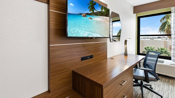 A modern hotel room with a wooden desk, a flat-screen TV mounted on the wall, a comfortable office chair, and a large window offering a view of palm trees and a beach. - Holiday Inn Express Harlingen by IHG (Harlingen)