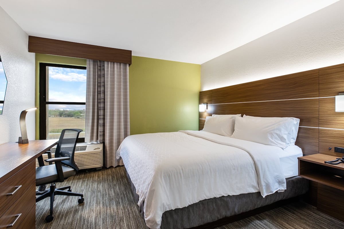 A hotel room with a large bed, a desk with a chair, a window with curtains, and a view of the outdoors.