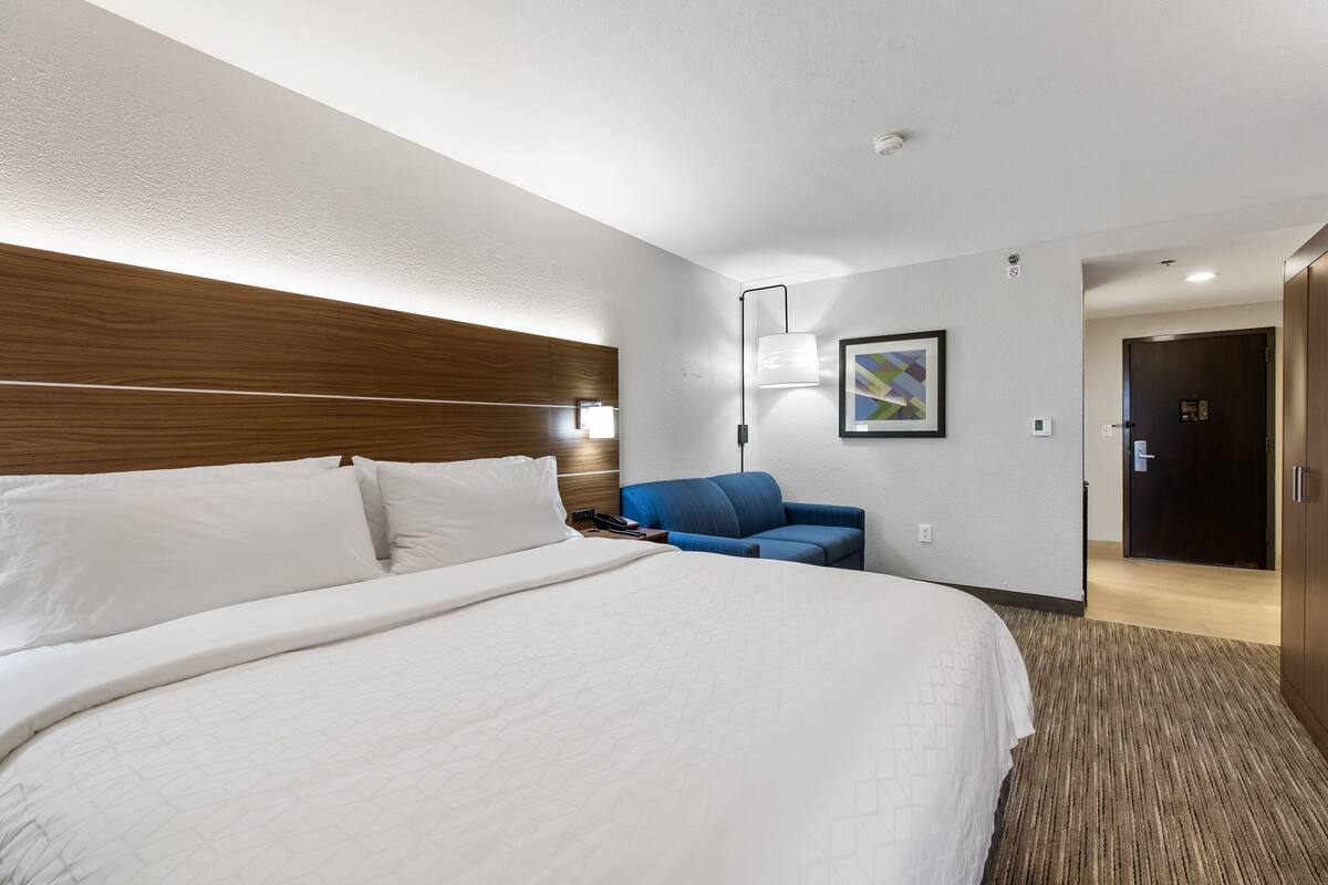 A hotel room with a large bed, a blue armchair, a desk, and a door.