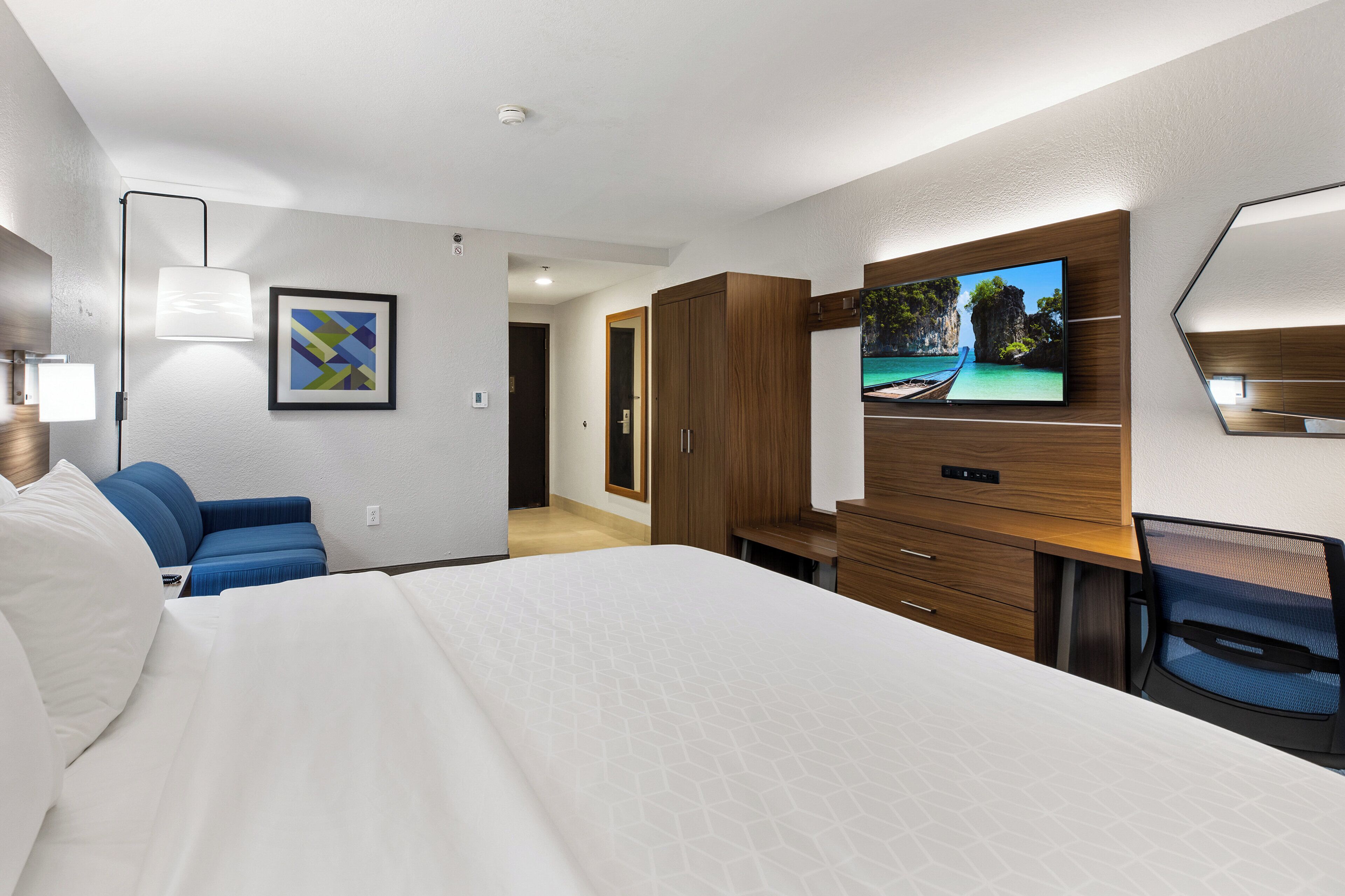 A hotel room with a large bed, a TV mounted on the wall, a desk, and a blue chair.