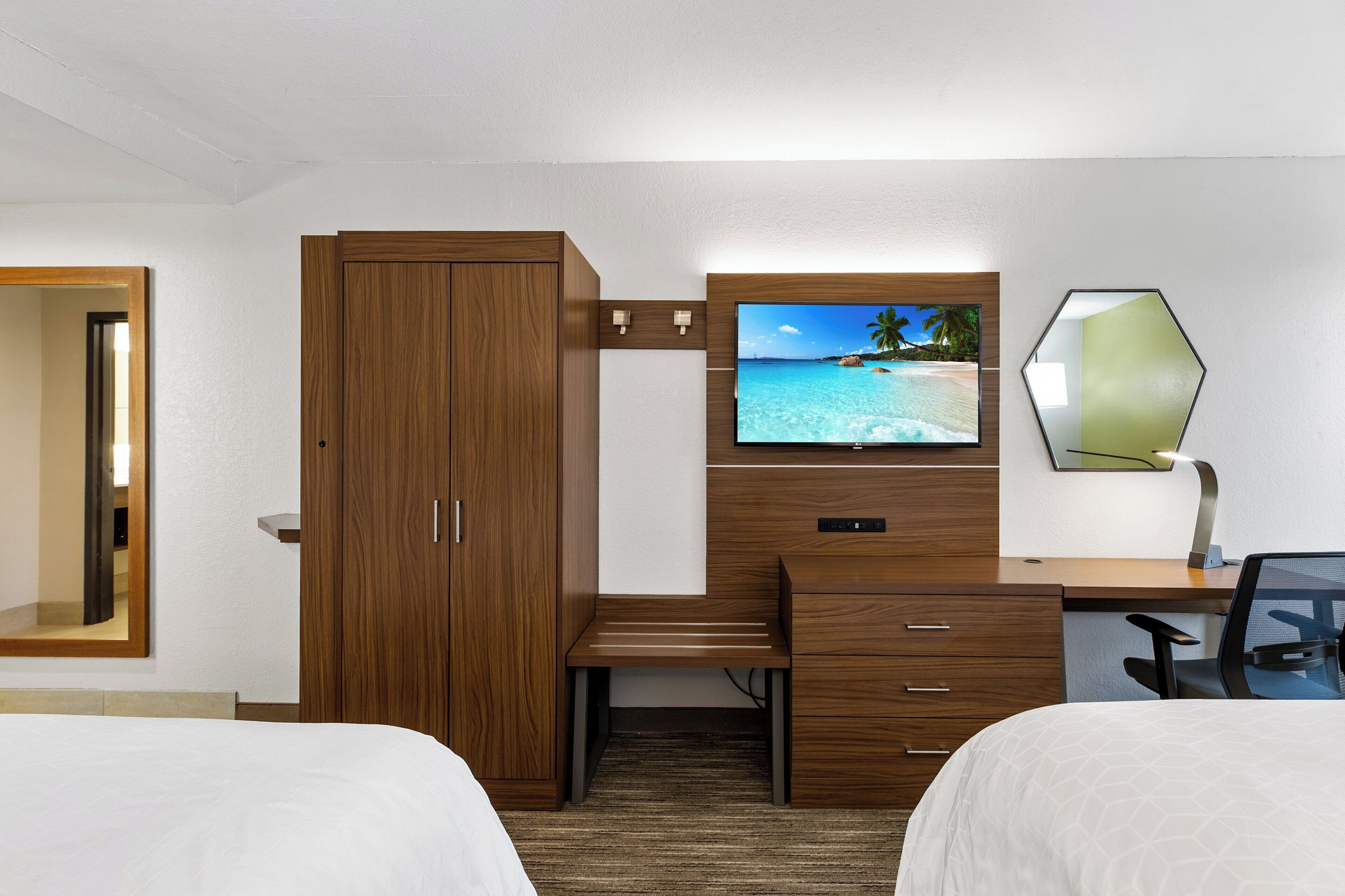A hotel room with a wooden desk, a television, a mirror, two beds, and a carpeted floor.
