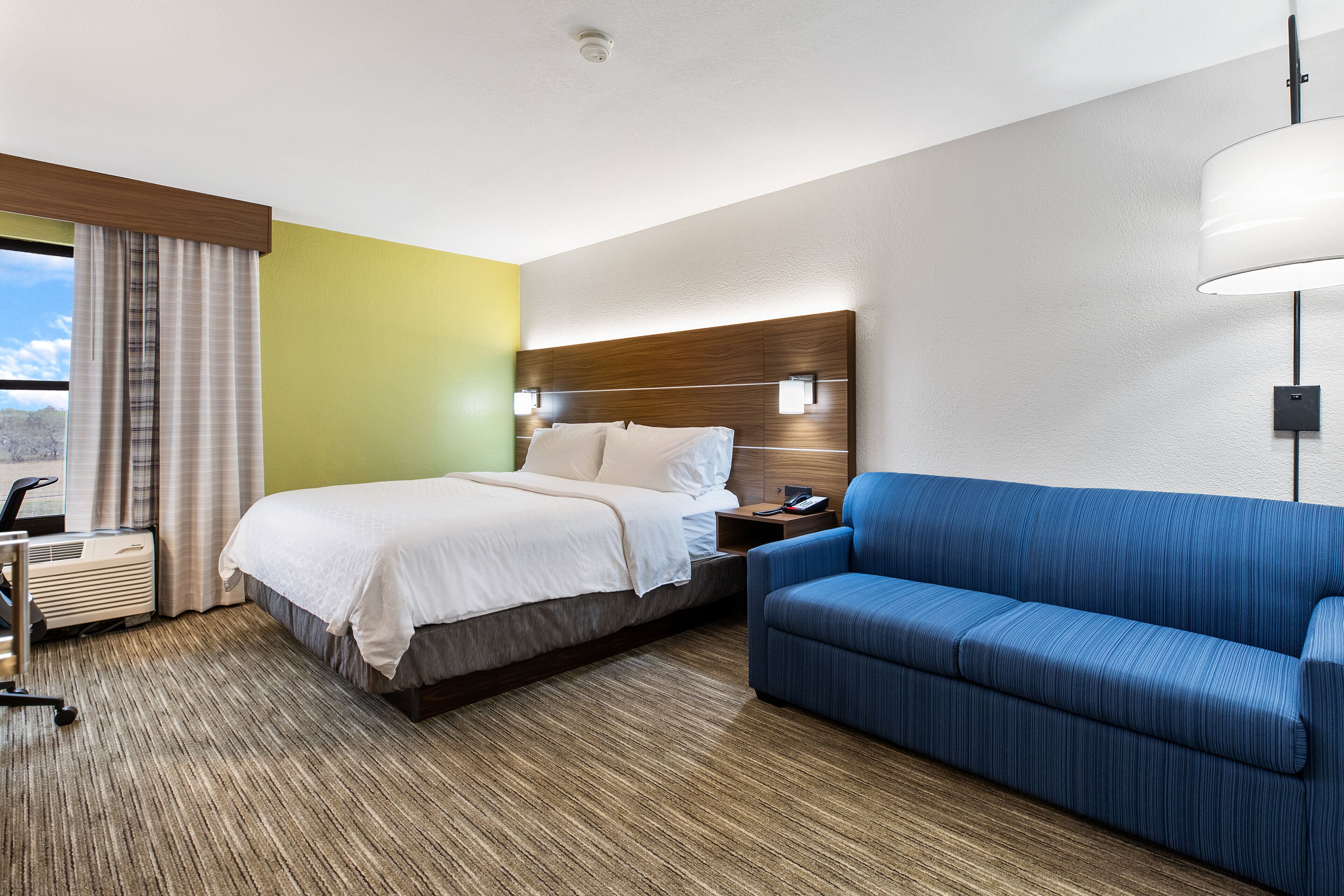 Habitación de hotel con una cama, un sofá azul, una mesita de noche con lámpara y una ventana con cortinas.