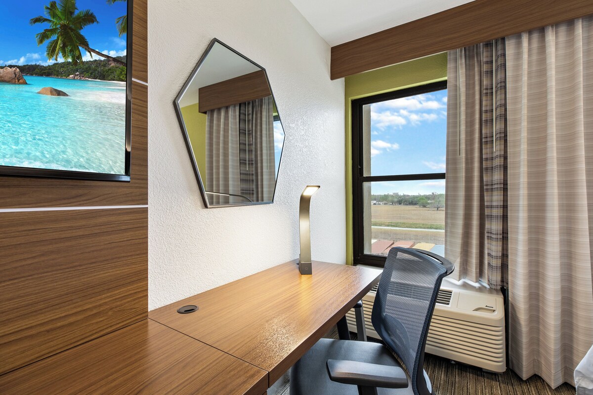 A hotel room with a wooden desk, a chair, a large mirror, a window with curtains, and a view of a beach.